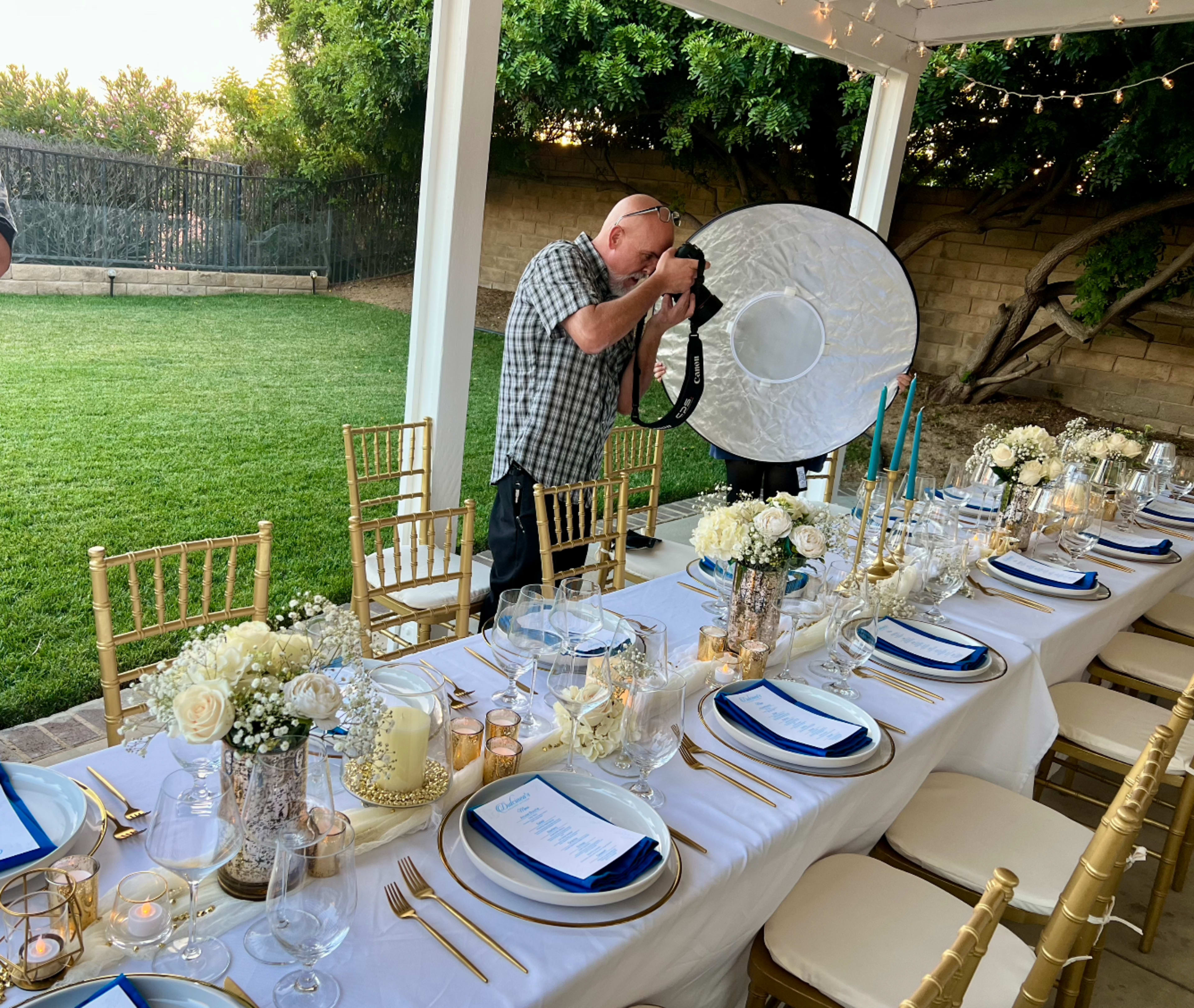 A photographer adjusts lighting equipment while capturing a beautifully set outdoor dining table adorned with white florals and elegant tableware.