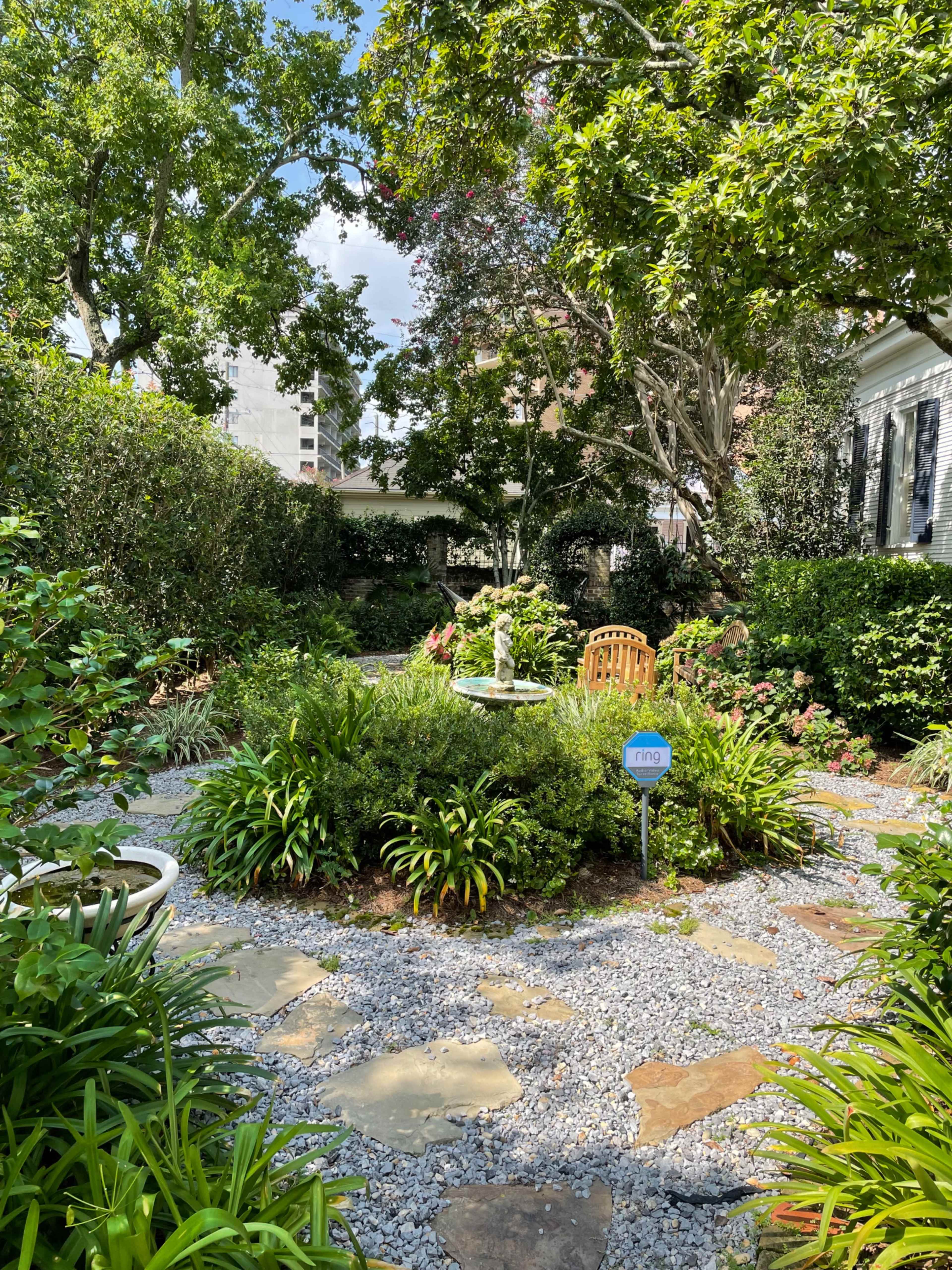 The scene features a landscaped garden with a central fountain surrounded by various plants and a pathway made of stone and gravel.