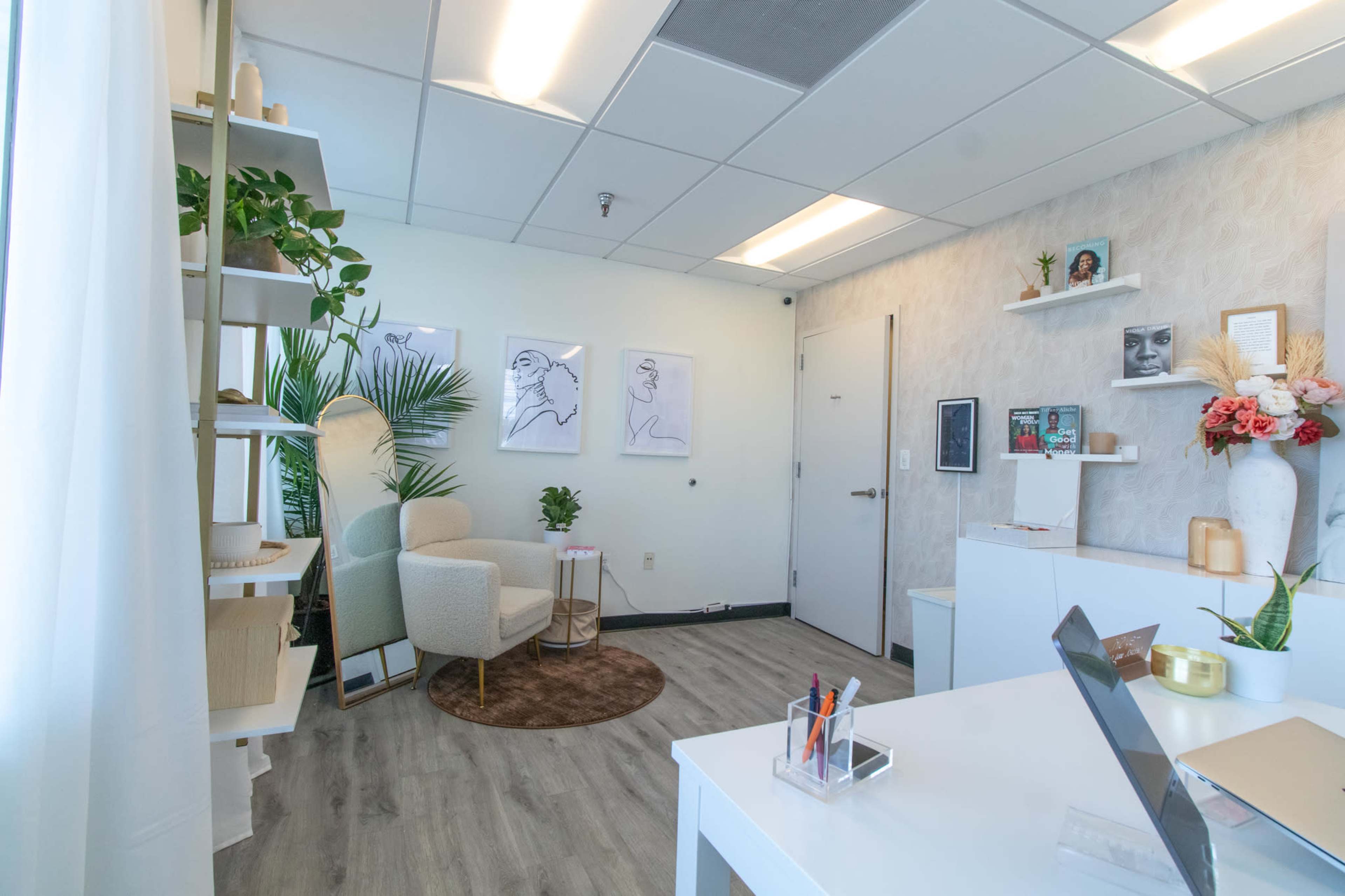 A bright, modern office space features a stylish seating area, plants, wall art, and a sleek desk with a laptop.