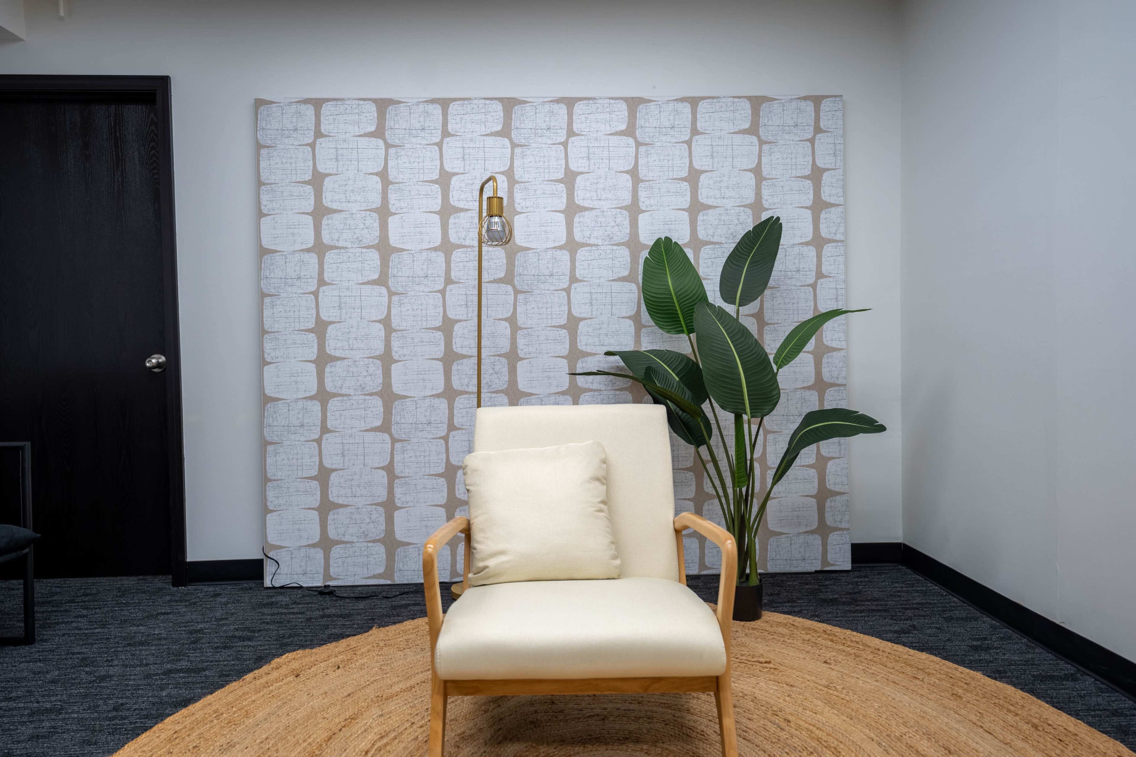 A light-colored chair sits on a circular rug in front of a patterned backdrop, alongside a tall plant and a floor lamp.