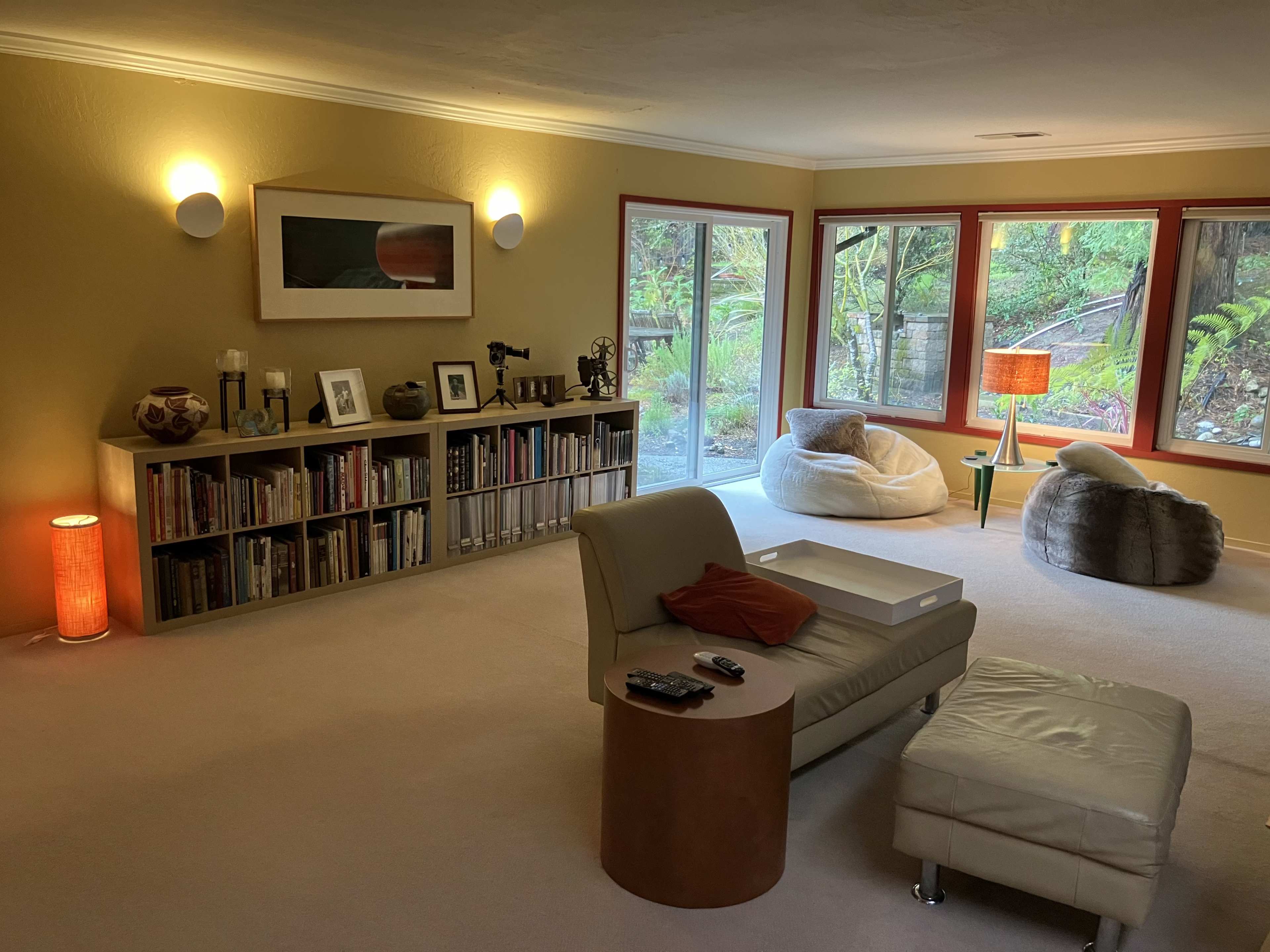 The room features a beige carpet, a bookshelf filled with books, a comfortable seating area with modern furniture, and large windows showcasing a green outdoor space.