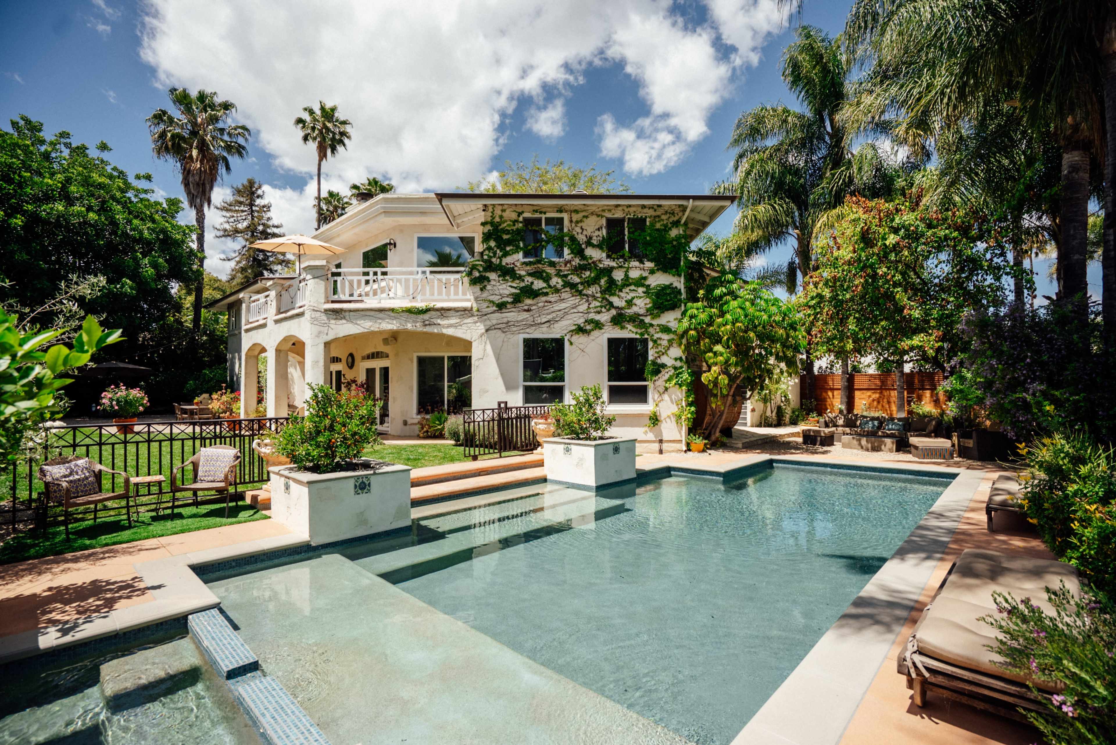 The image shows a two-story house surrounded by green landscaping and a rectangular swimming pool in the foreground.
