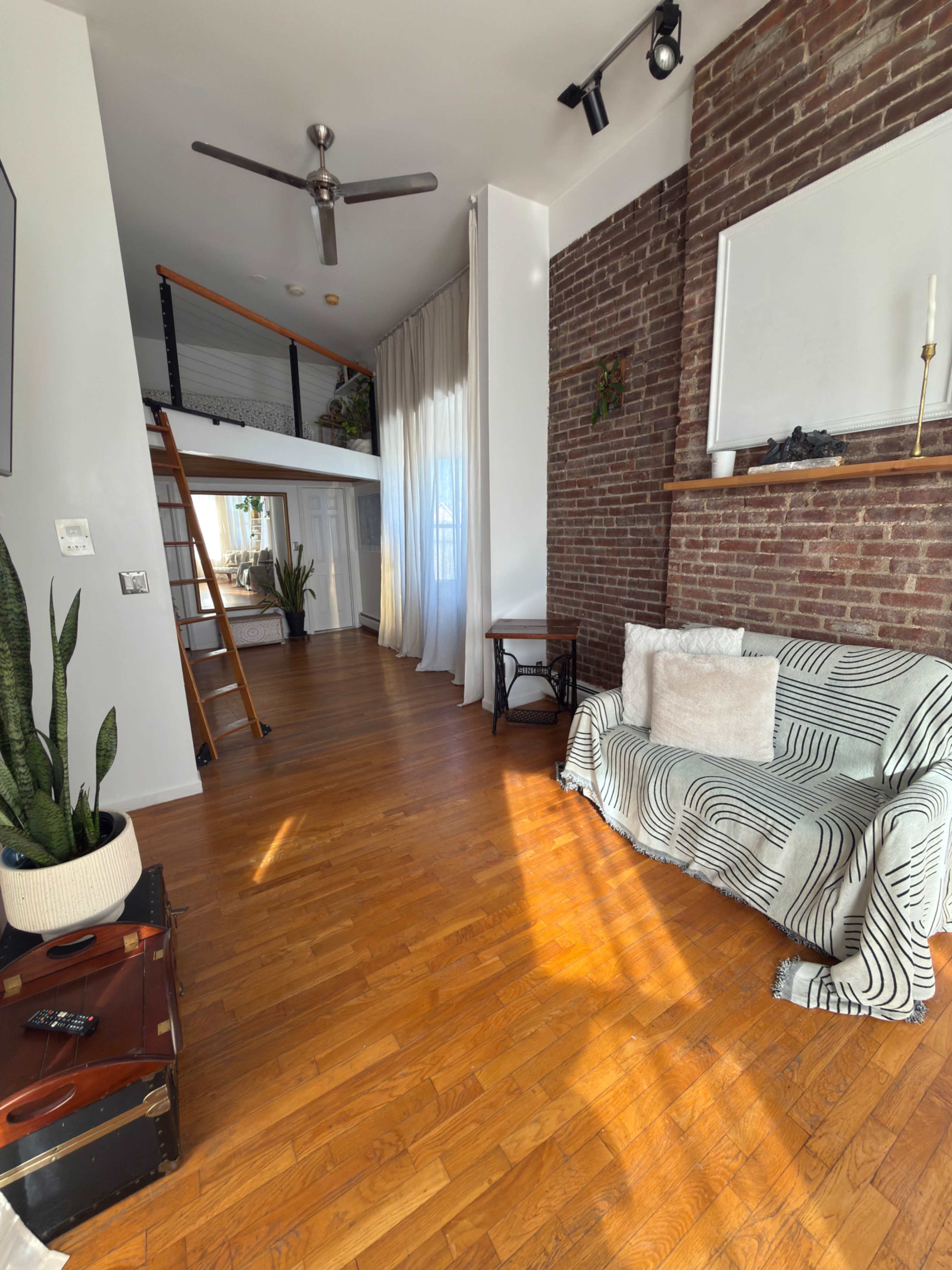 The image shows a spacious living area with hardwood floors, a brick wall, a white sofa with a patterned throw, a ladder leading to an upper level, and large windows with sheer curtains.