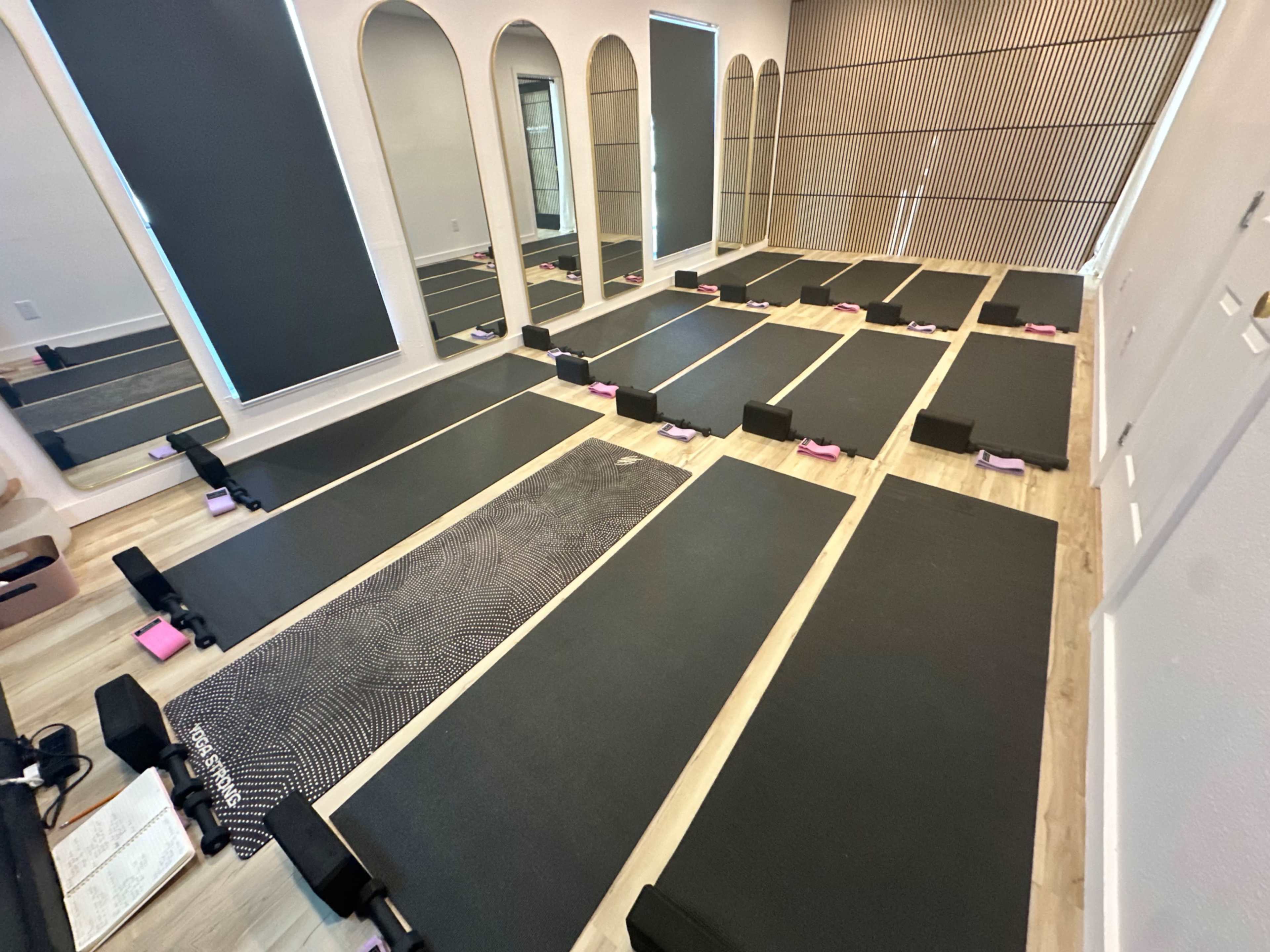 The image shows a yoga or fitness studio with several exercise mats arranged in rows on a wooden floor, surrounded by mirrors and partial wall panels.