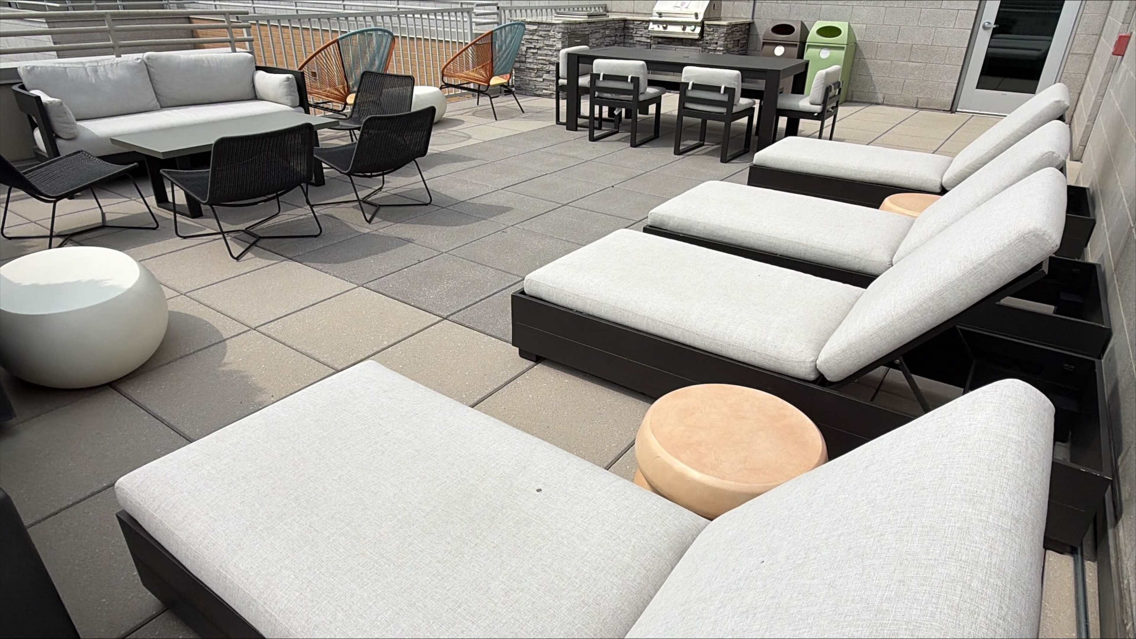 The image shows a rooftop terrace featuring modern lounge chairs, a dining table, and a barbecue area surrounded by a concrete wall.