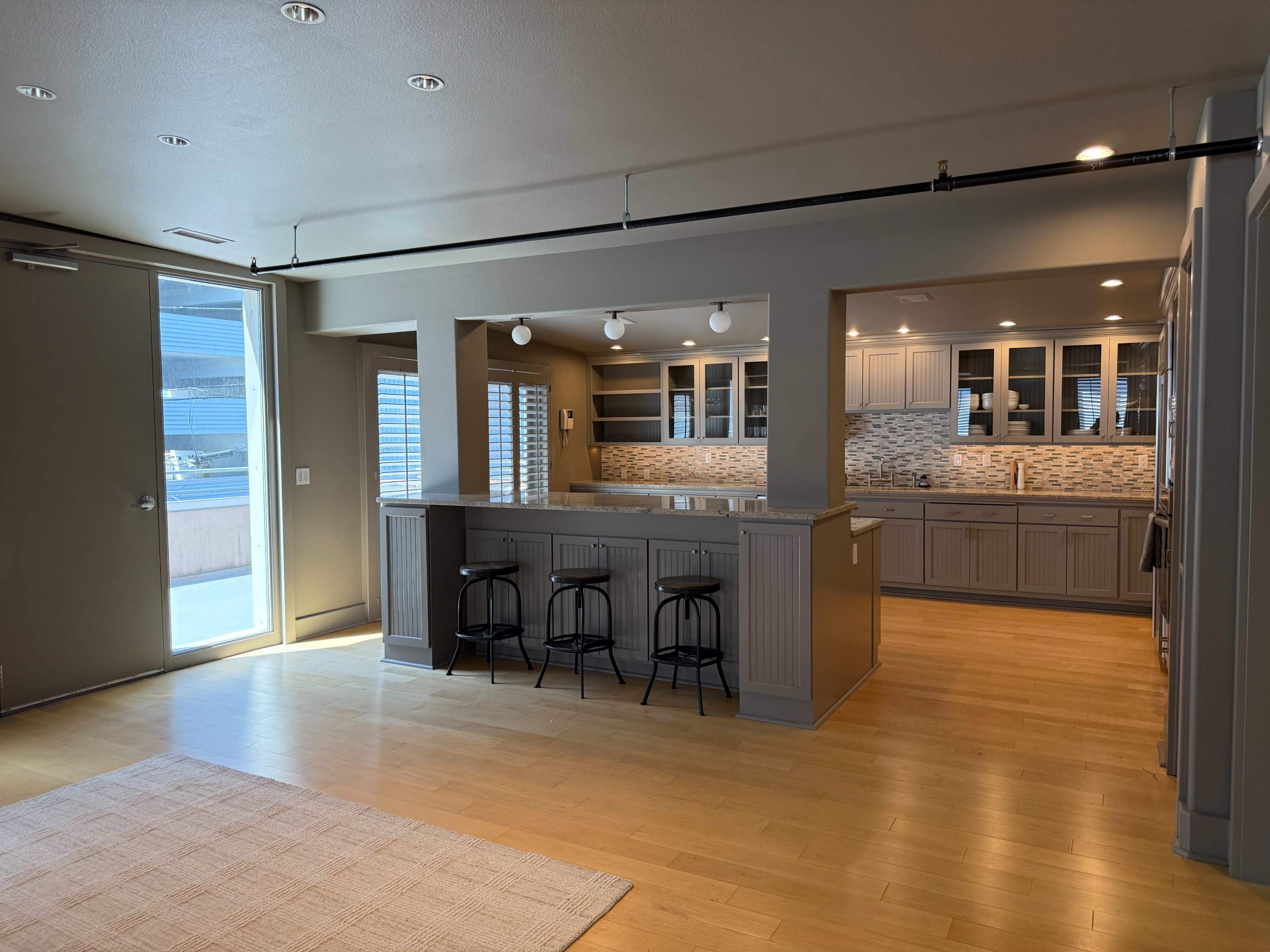 The image shows a modern kitchen and living area with wooden flooring, a large island with bar stools, and ample cabinetry.