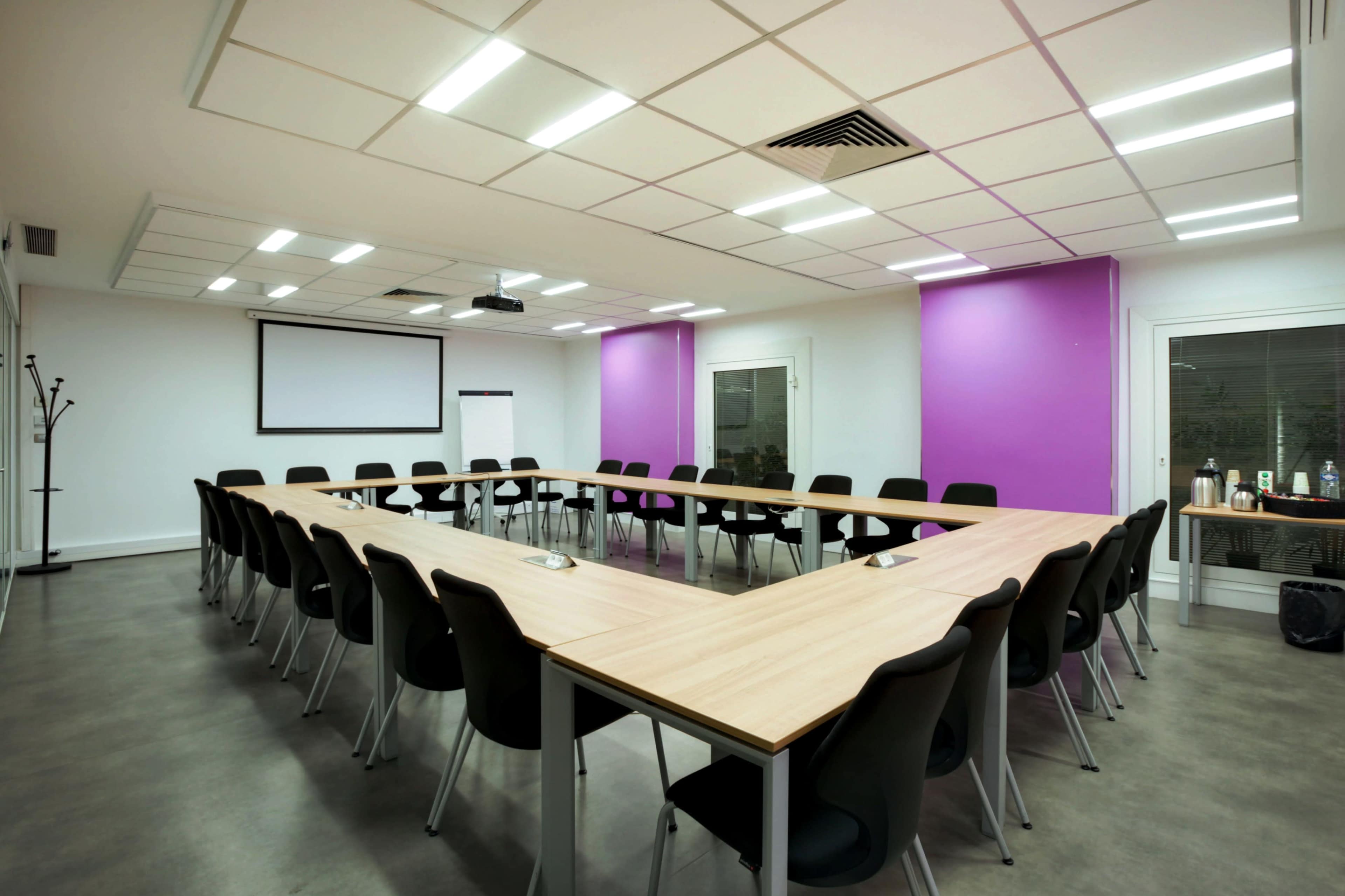A conference room features a long, triangular table surrounded by black chairs, with a projector screen at the front and purple accent walls.