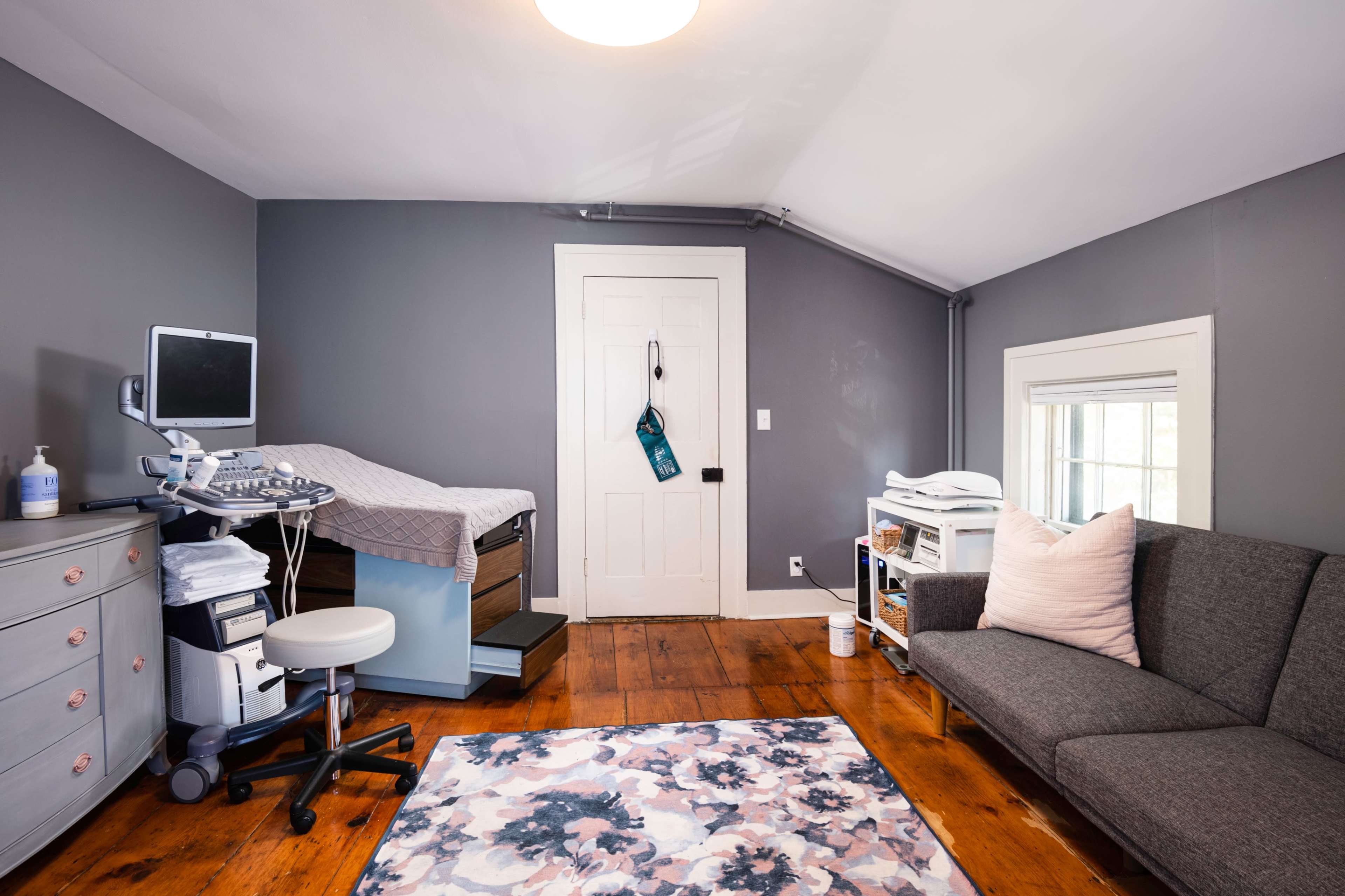 A small room with a medical examination table, a desk with medical equipment, and a sofa, featuring wooden floors and a window.