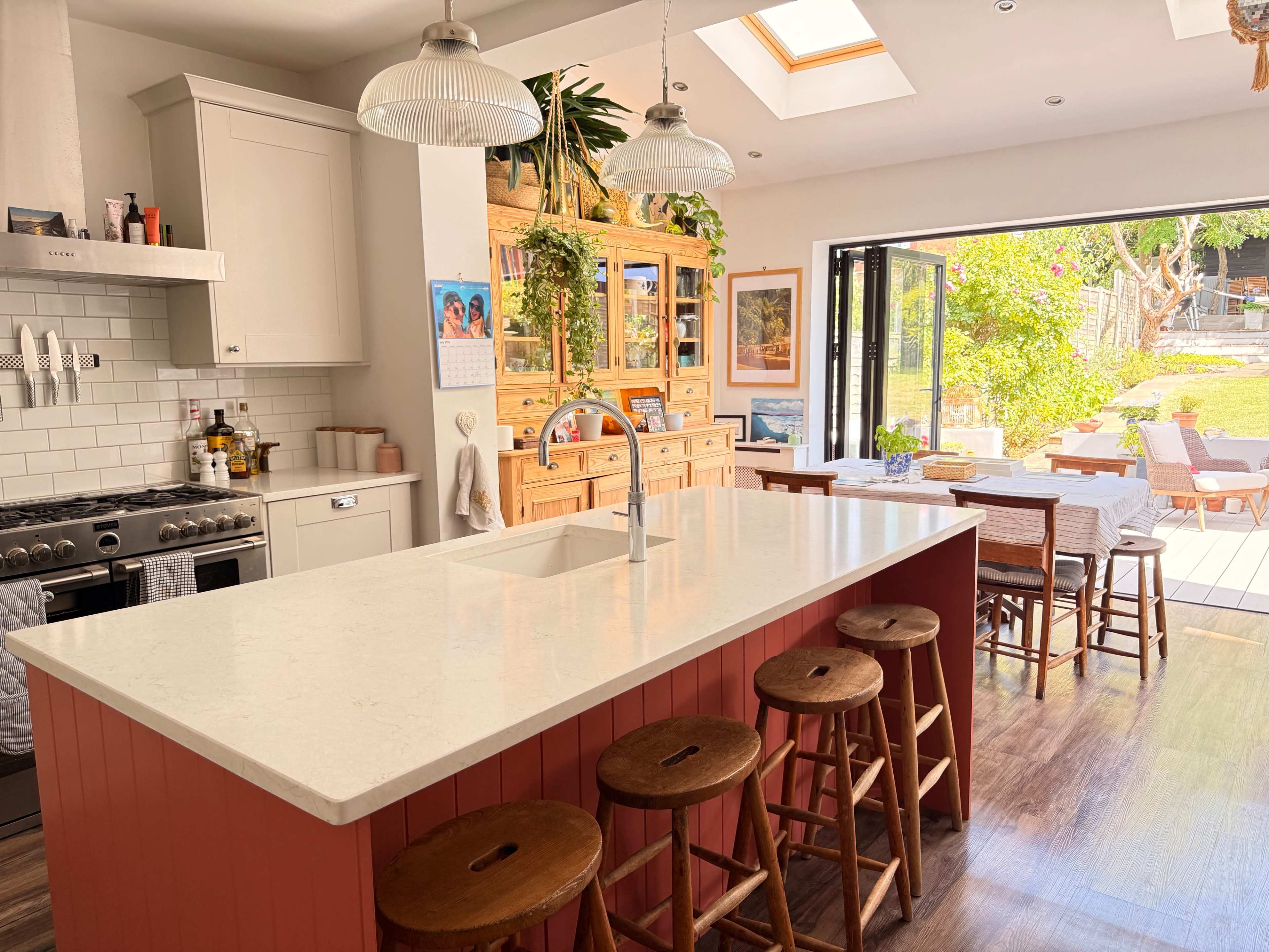 The image shows a modern kitchen with a large central island, bar stools, wooden cabinetry, and an open layout leading to an outdoor dining area.