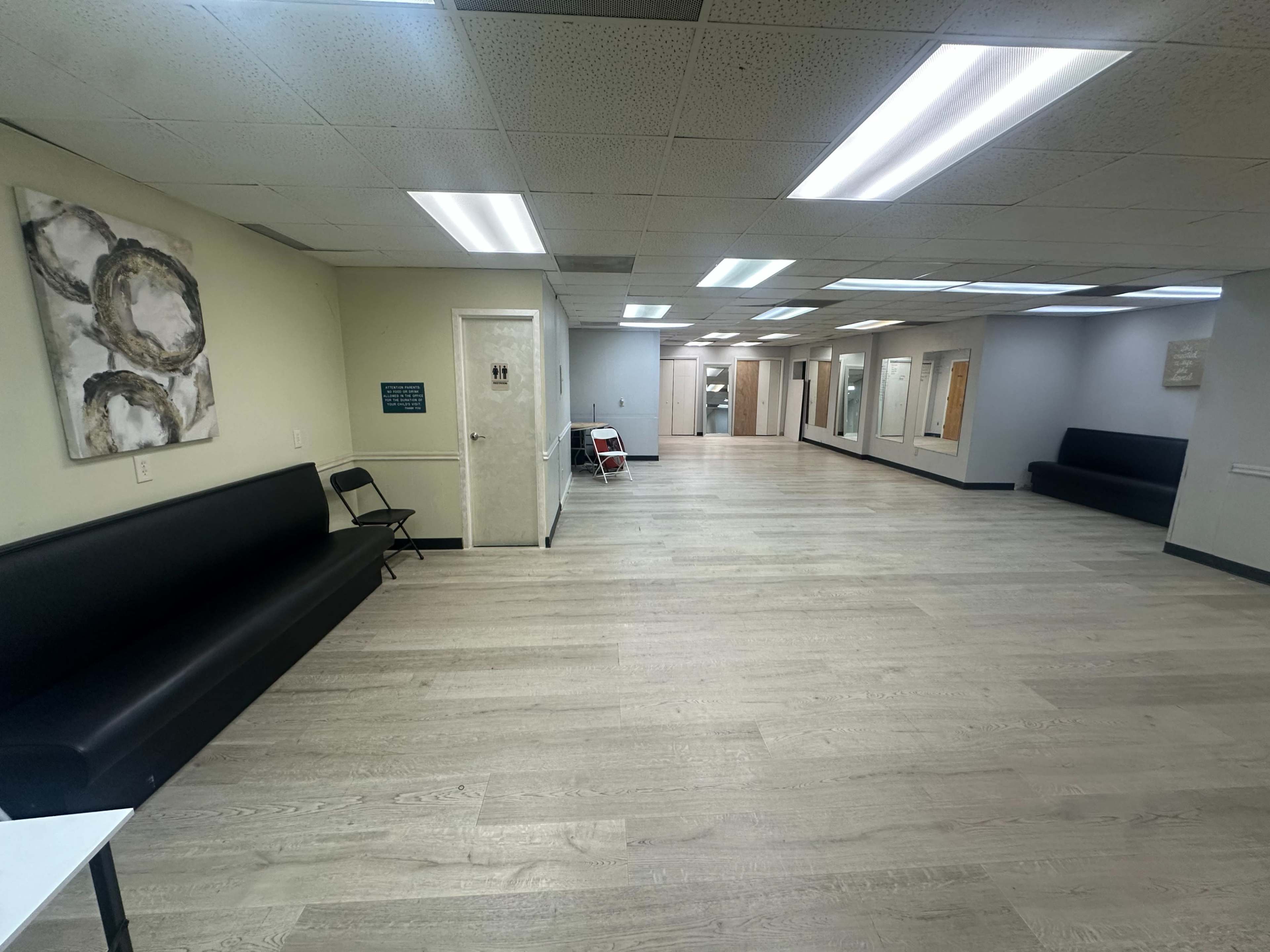 The image shows a spacious waiting area with wooden flooring, a few benches along the walls, and several doors leading to other rooms in a well-lit facility.