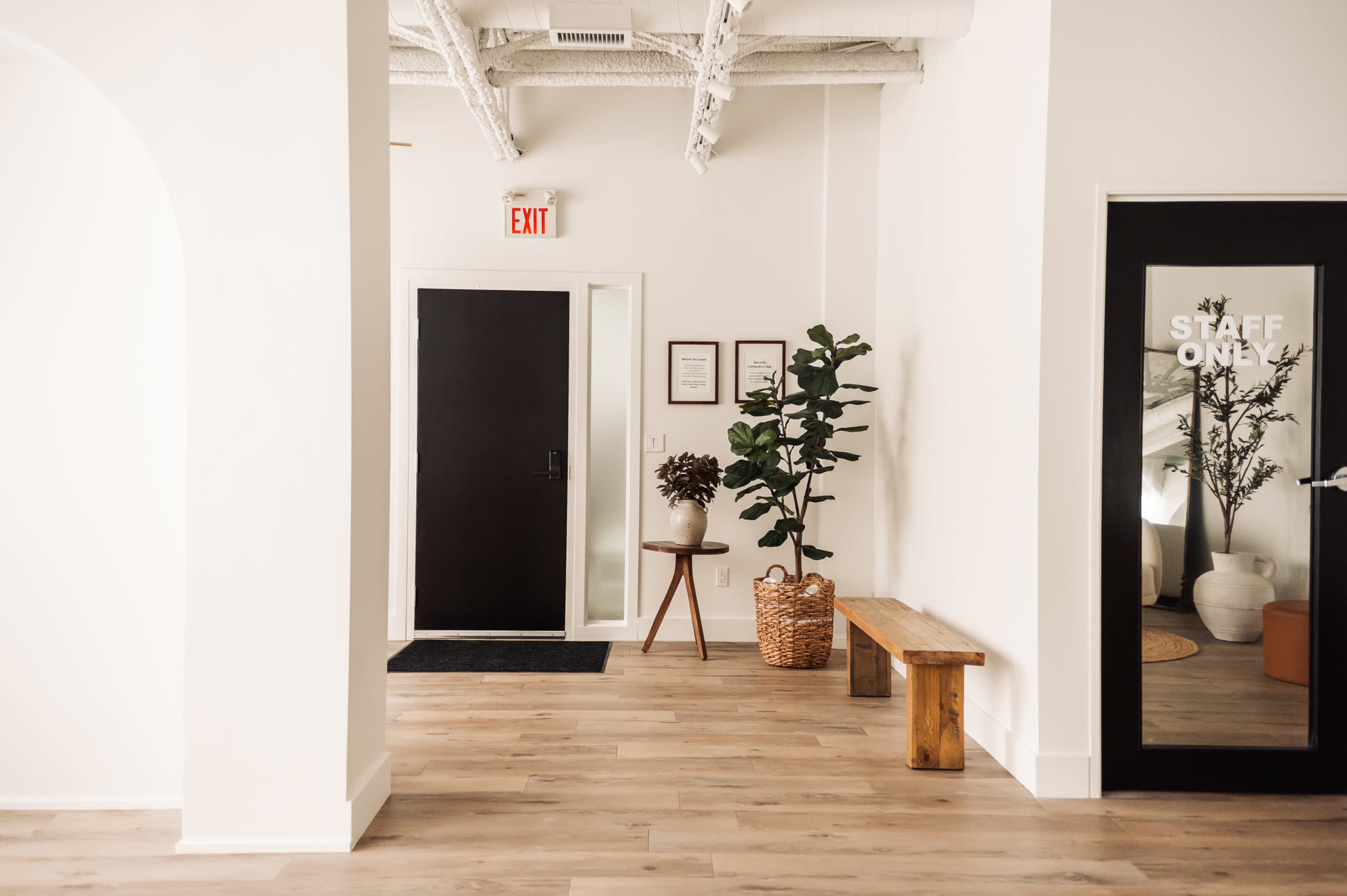 The image shows a well-lit hallway with a black door, a bench made of wood, a potted plant, and a glass door labeled "STAFF ONLY."