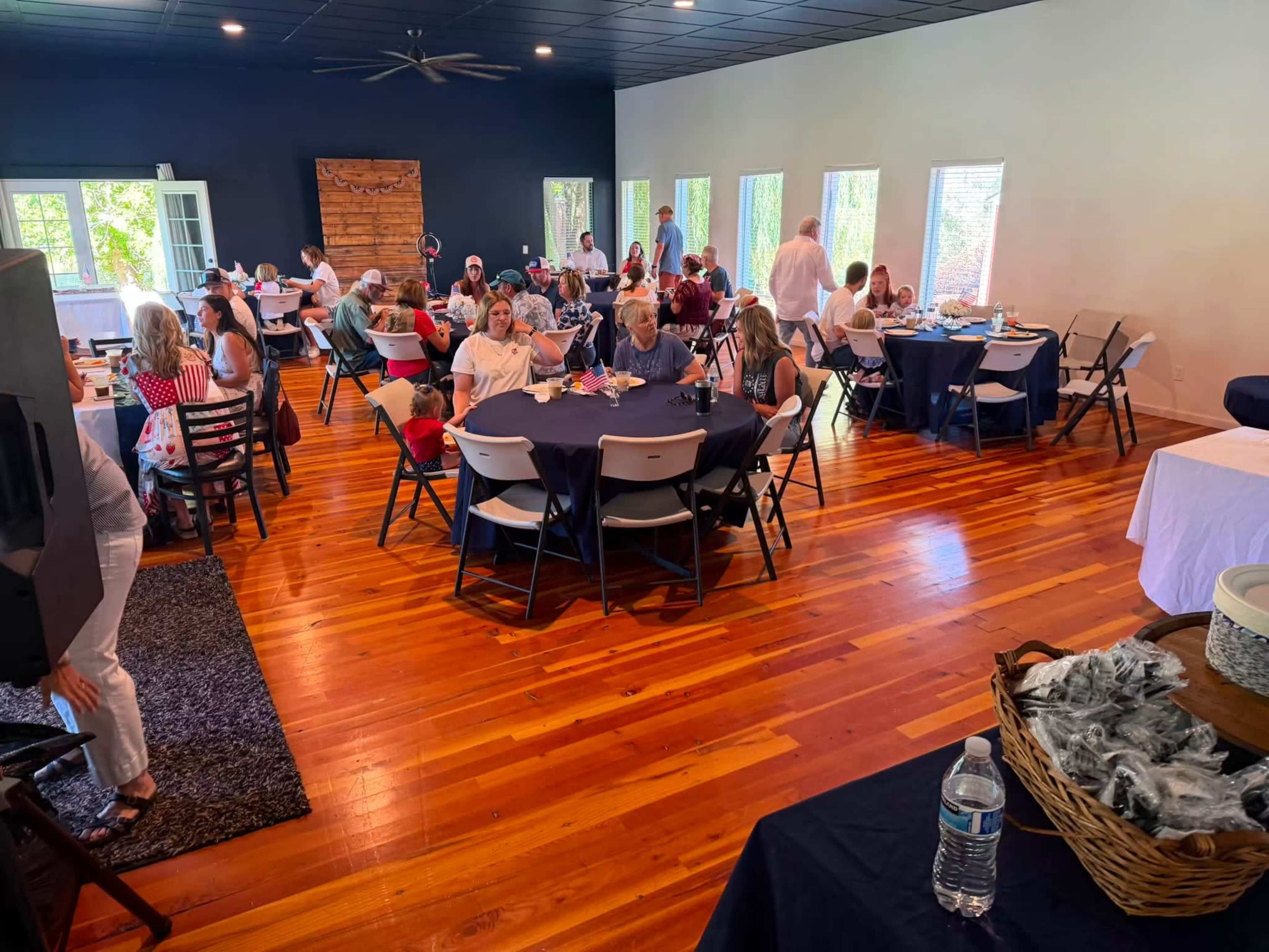 The image shows a gathering of people seated at tables in a well-lit banquet hall with wooden flooring and dark walls.