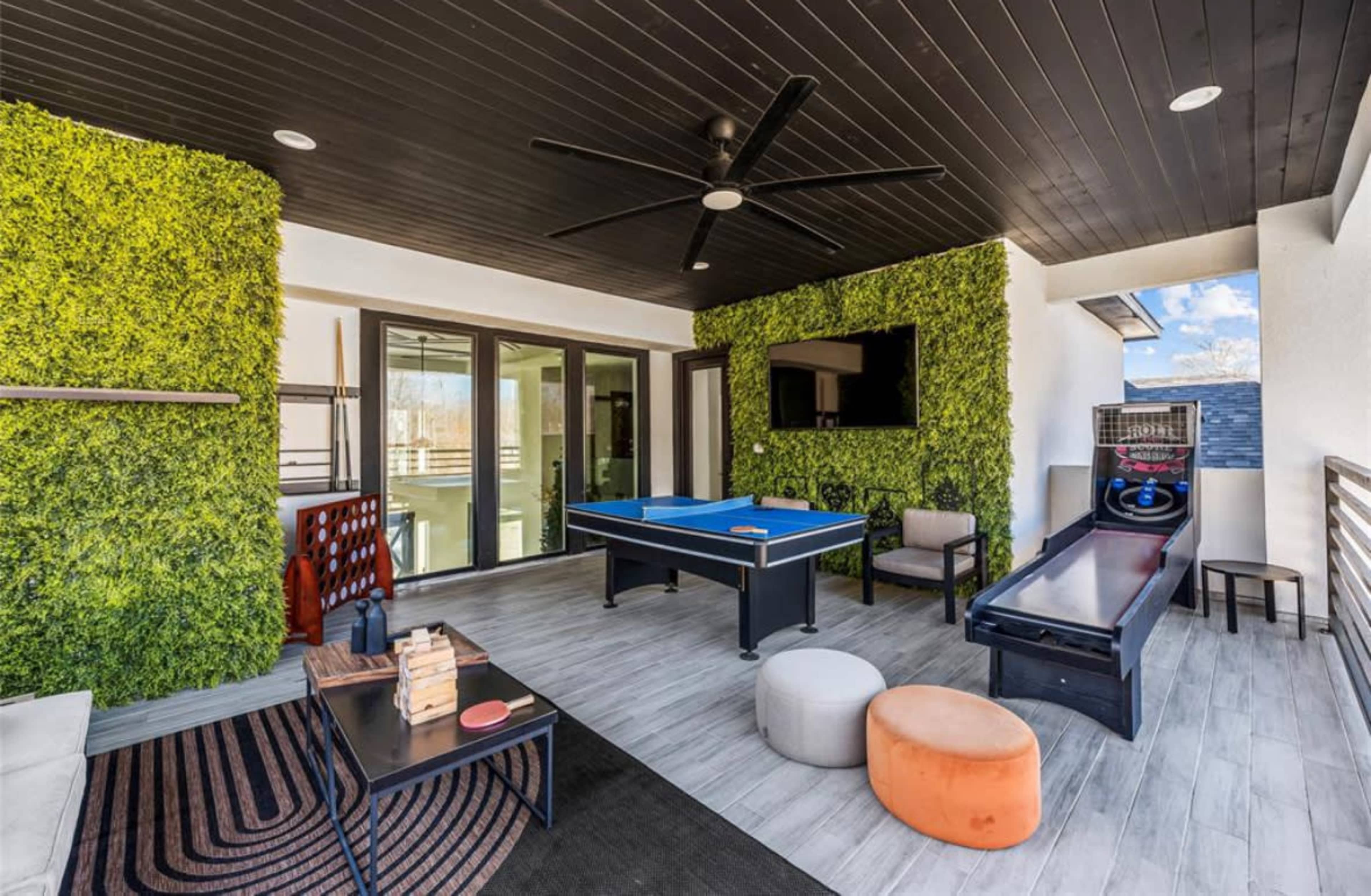 The image shows a modern outdoor patio featuring a pool table, a game arcade machine, seating areas, and a wall covered with greenery.