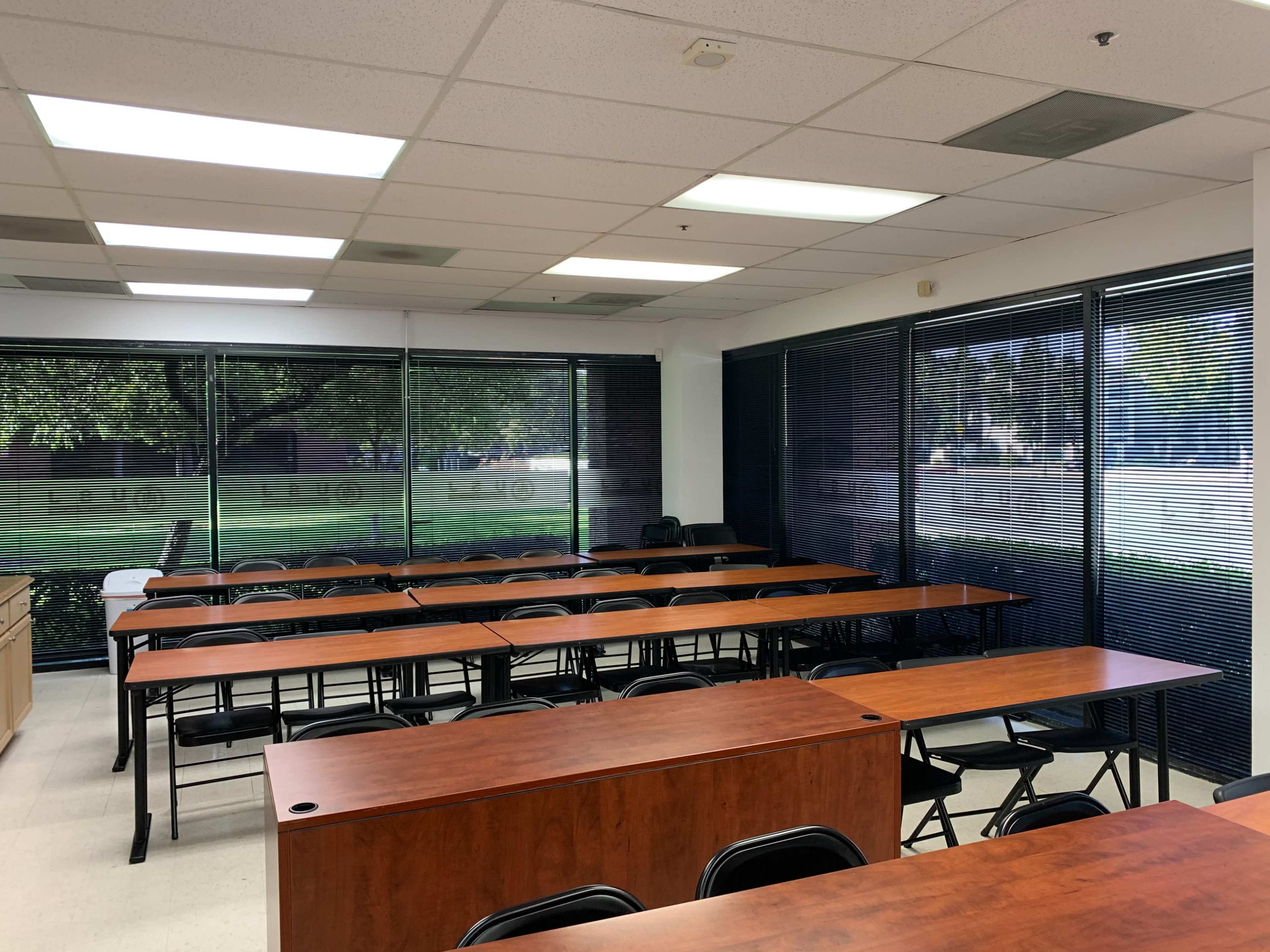 The image shows a classroom with rows of wooden desks and chairs arranged neatly, large windows on one side featuring blinds and a view of greenery outside.