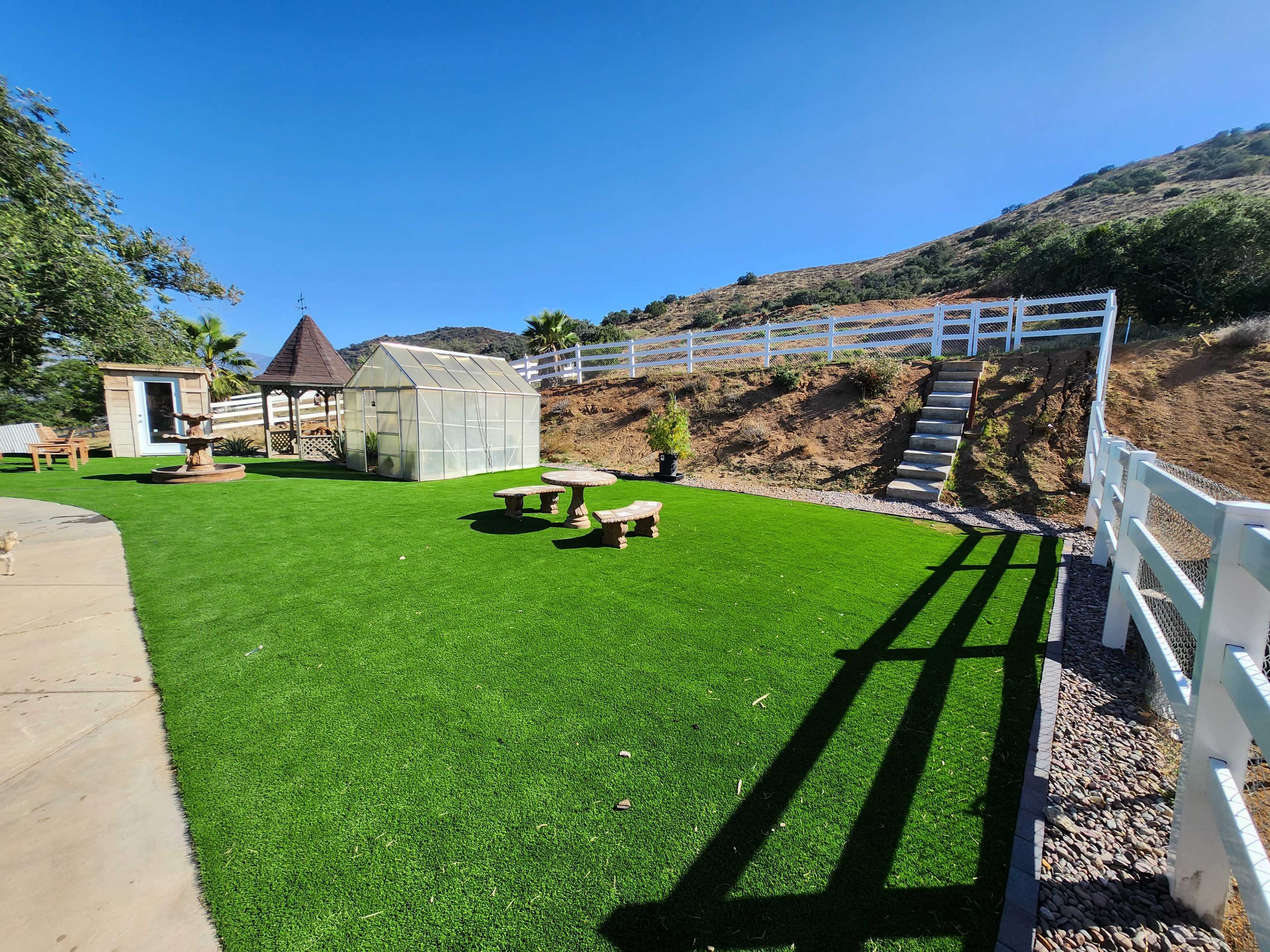A landscaped outdoor area with artificial grass, a greenhouse, benches, a water feature, and a hillside with a white fence.