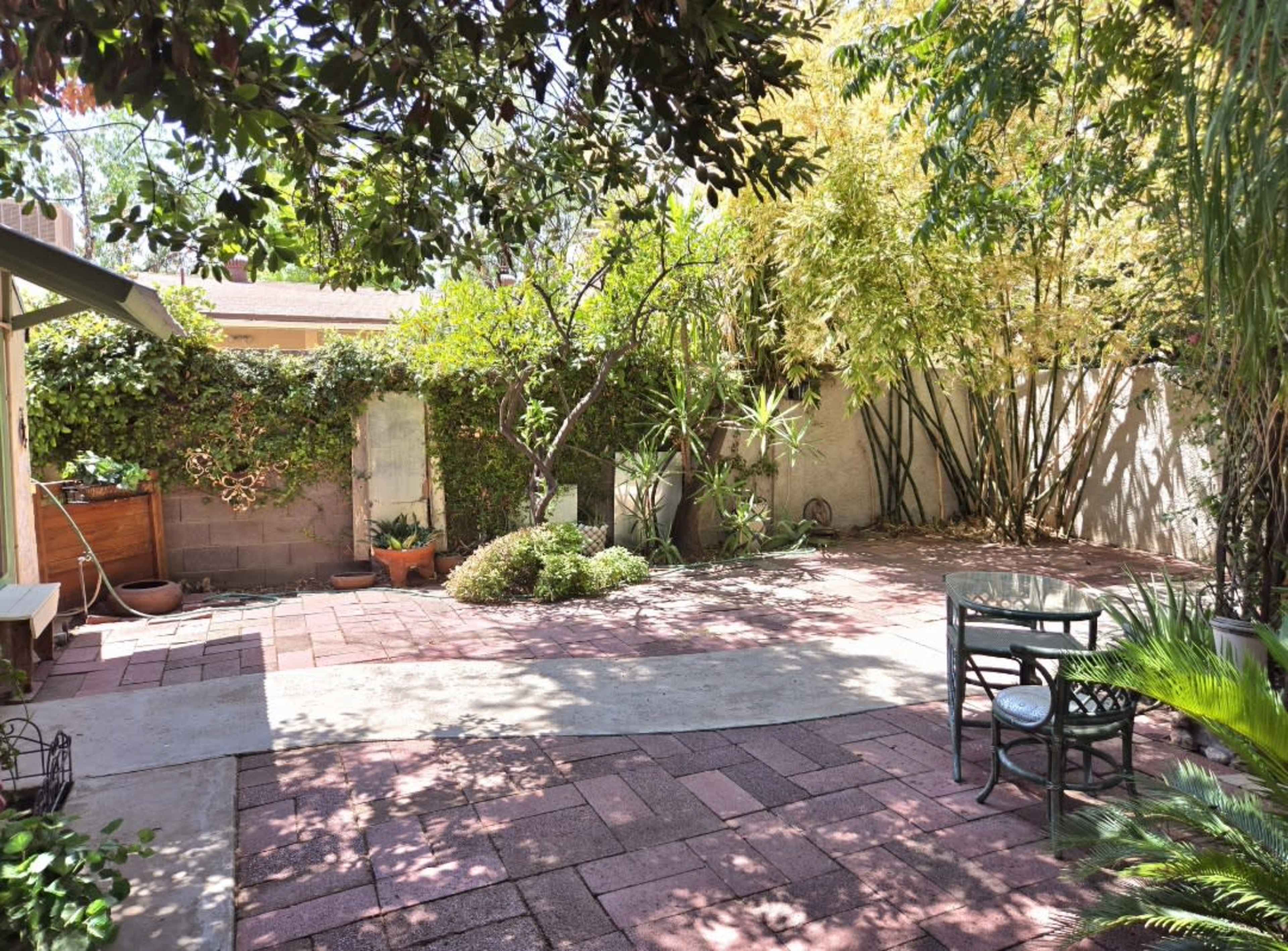 A flat, paved backyard features various plants and trees, with a table and chair set off to the side.