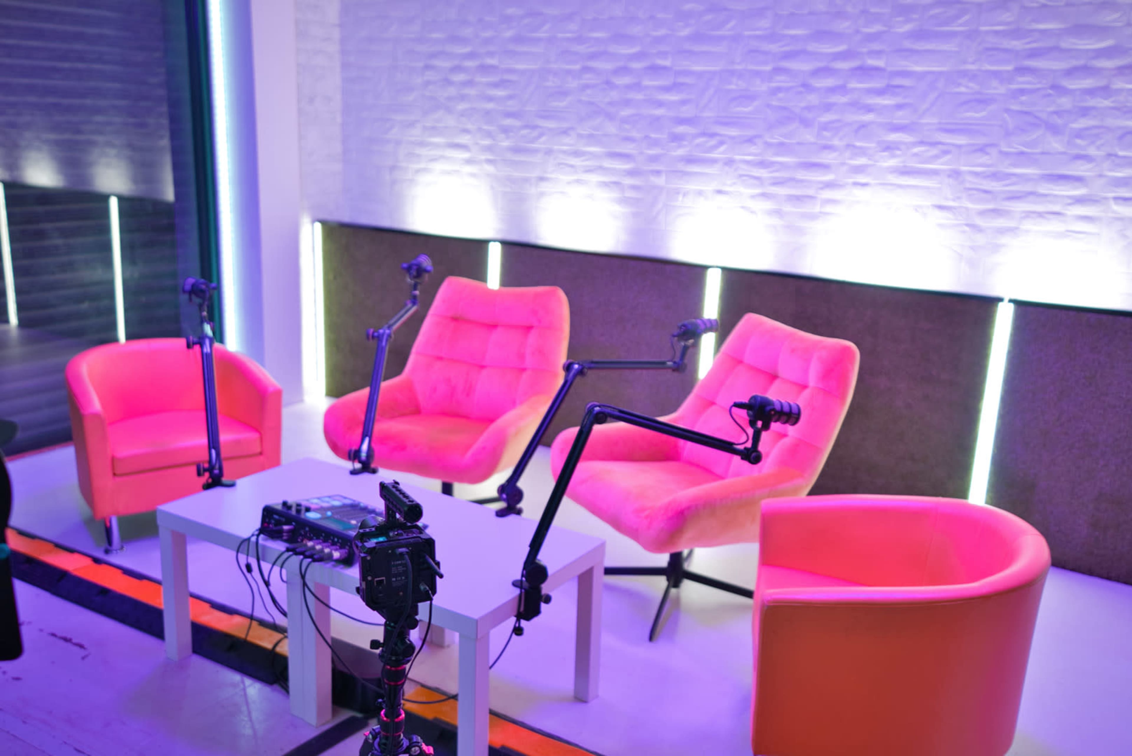 A modern studio setup with three pink chairs and a table, equipped with microphone arms and lighting.