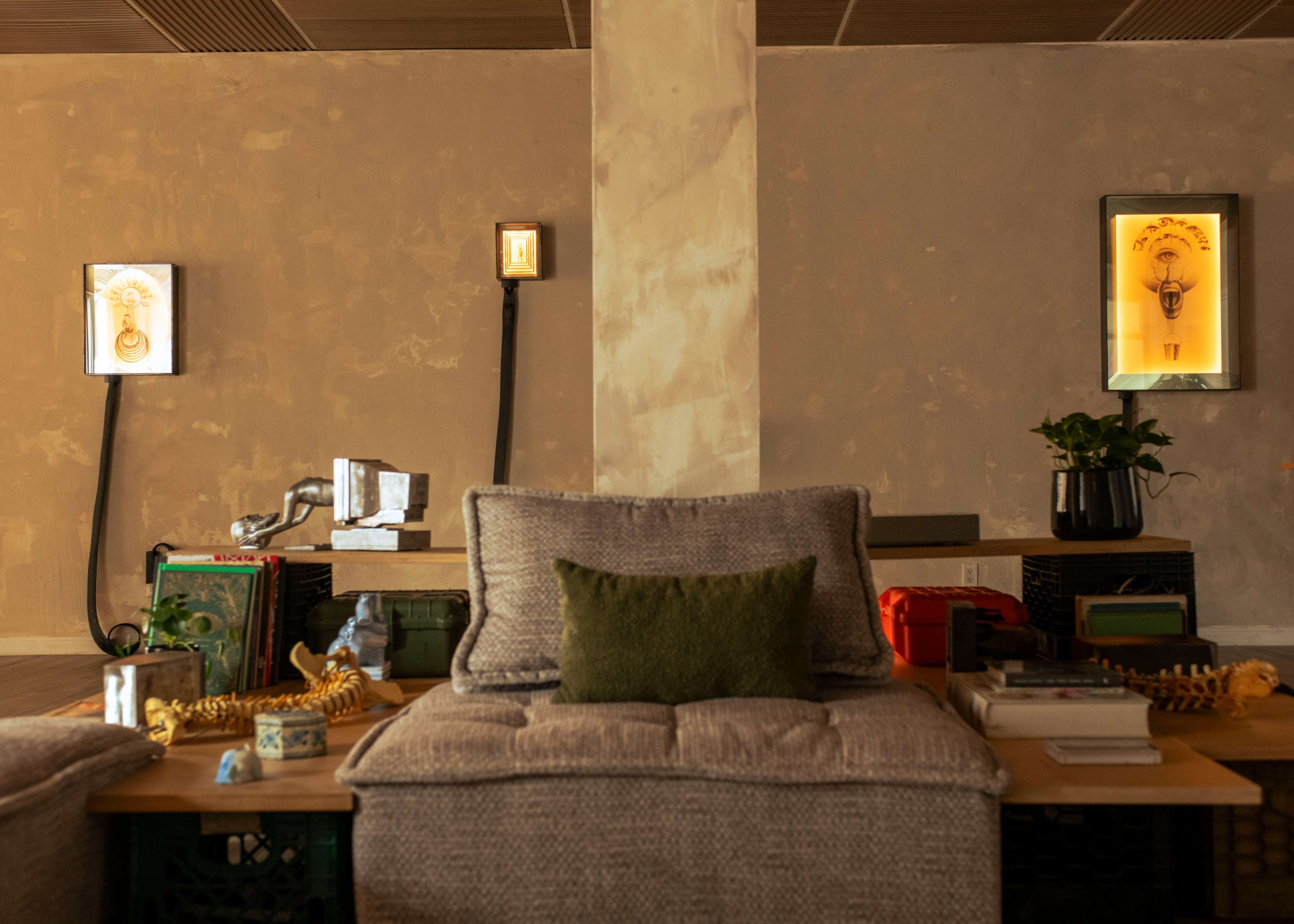 The image shows a cozy, minimalist living space with two wall-mounted light fixtures, a sofa adorned with cushions, and various decorative items on wooden shelves.