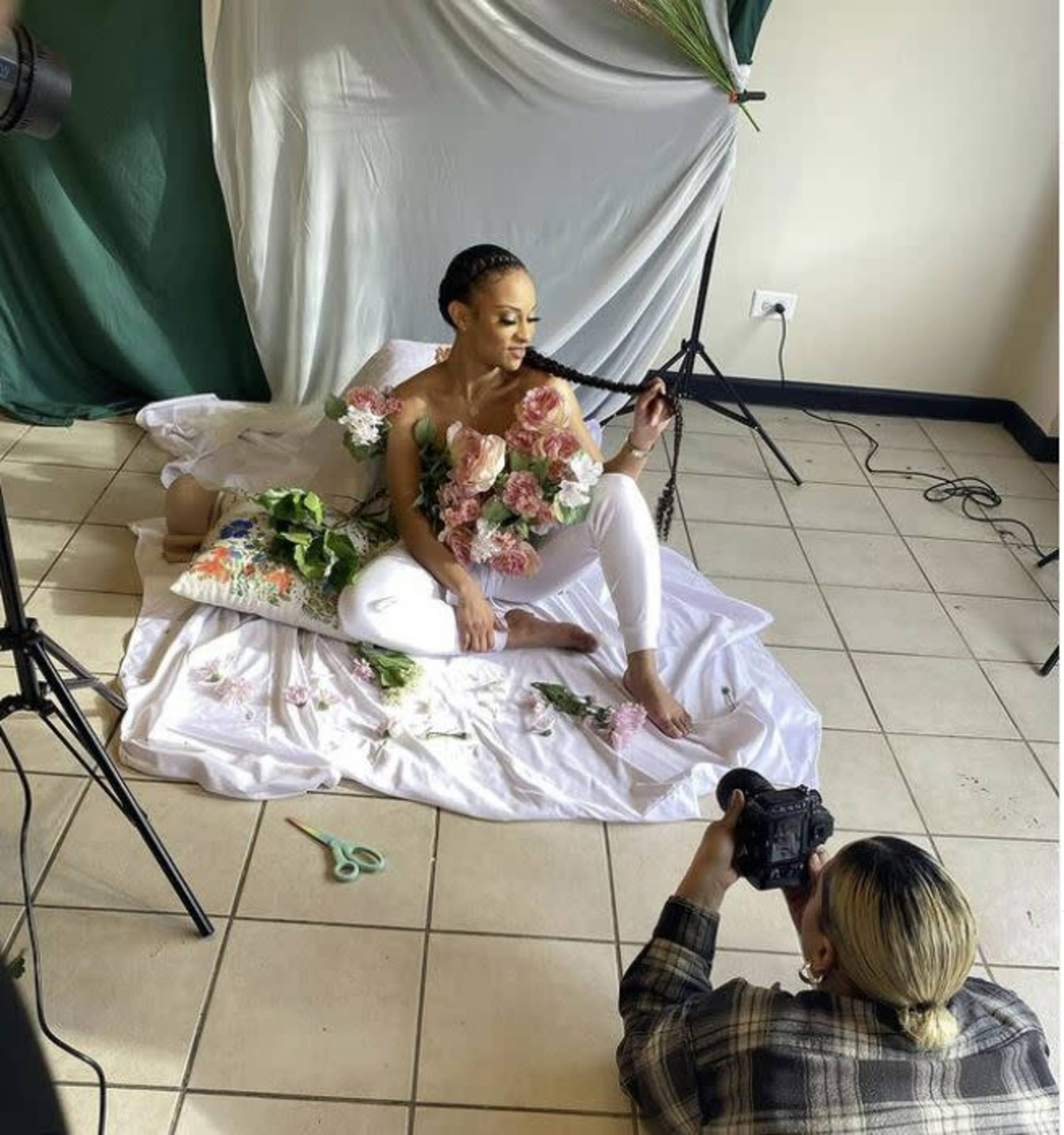 A model poses with flowers in her arms while a photographer captures the scene on a tiled floor in a studio setting.