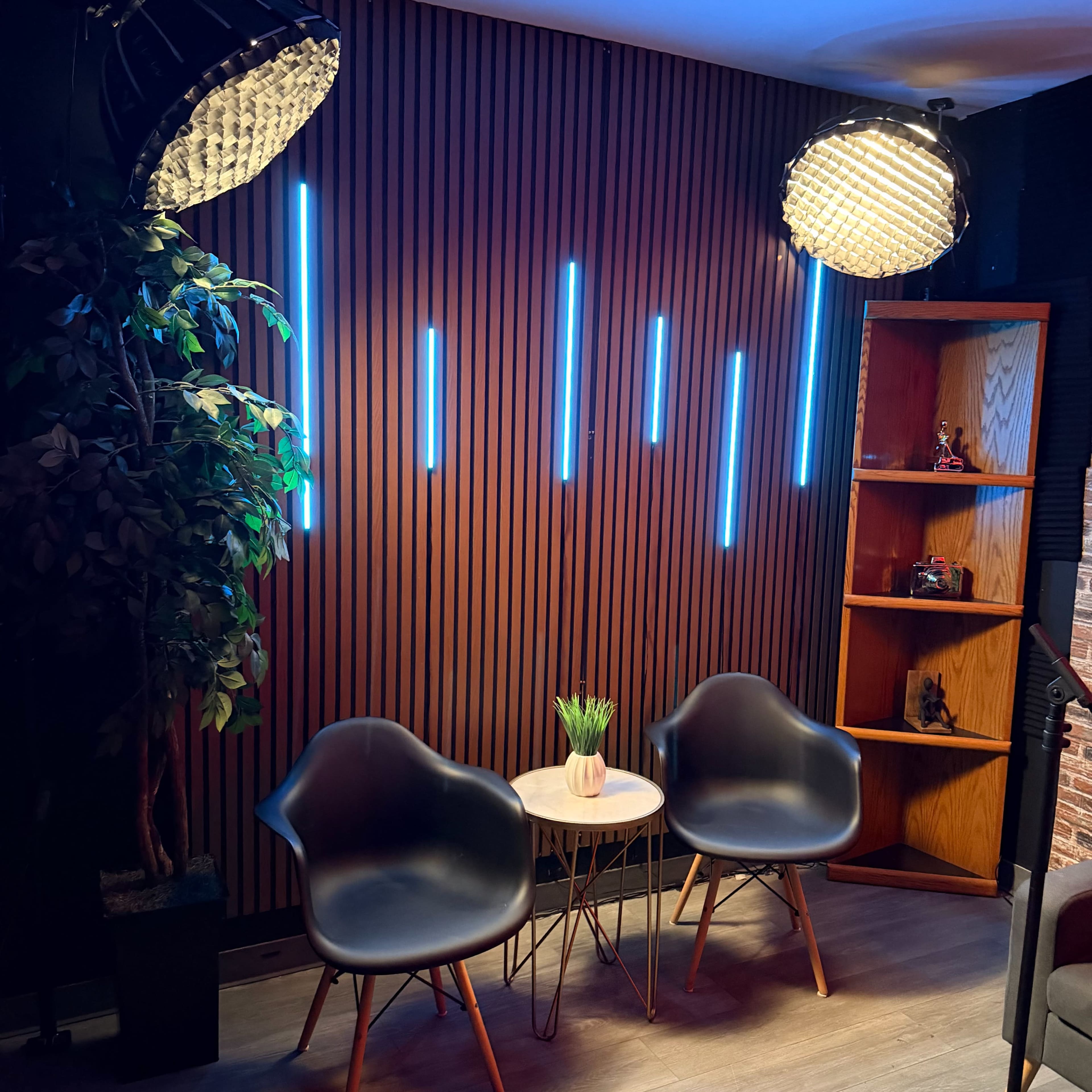 The image features a modern seating area with two black chairs, a small round table, and a wooden wall with vertical blue lighting, alongside a plant and a bookshelf.