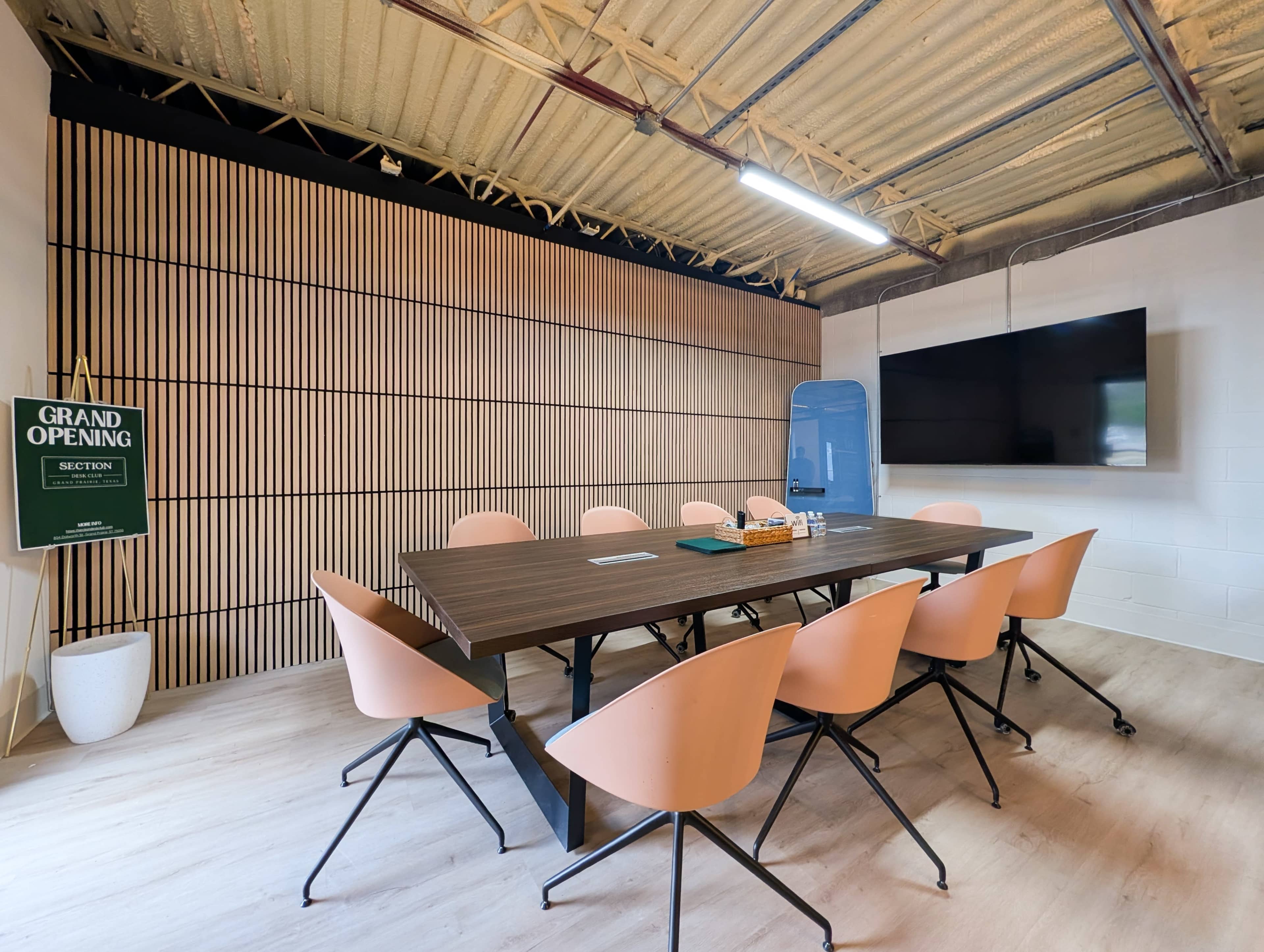 A modern conference room with a wooden table, eight chairs, a large television screen, and a "Grand Opening" sign on the wall.