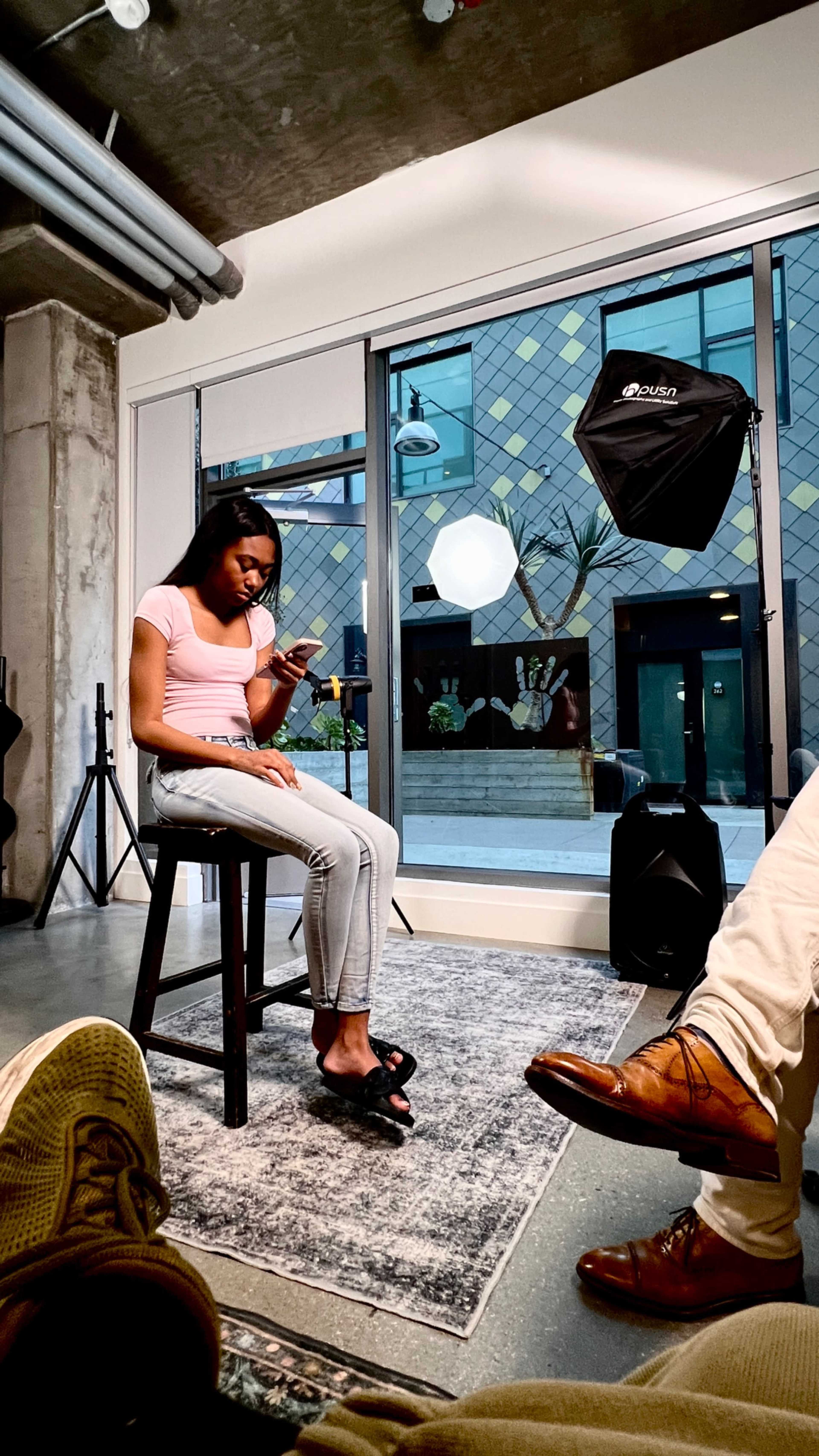 A young woman sits on a stool indoors, looking at her phone, with soft lighting and a large window showing an outdoor scene in the background.