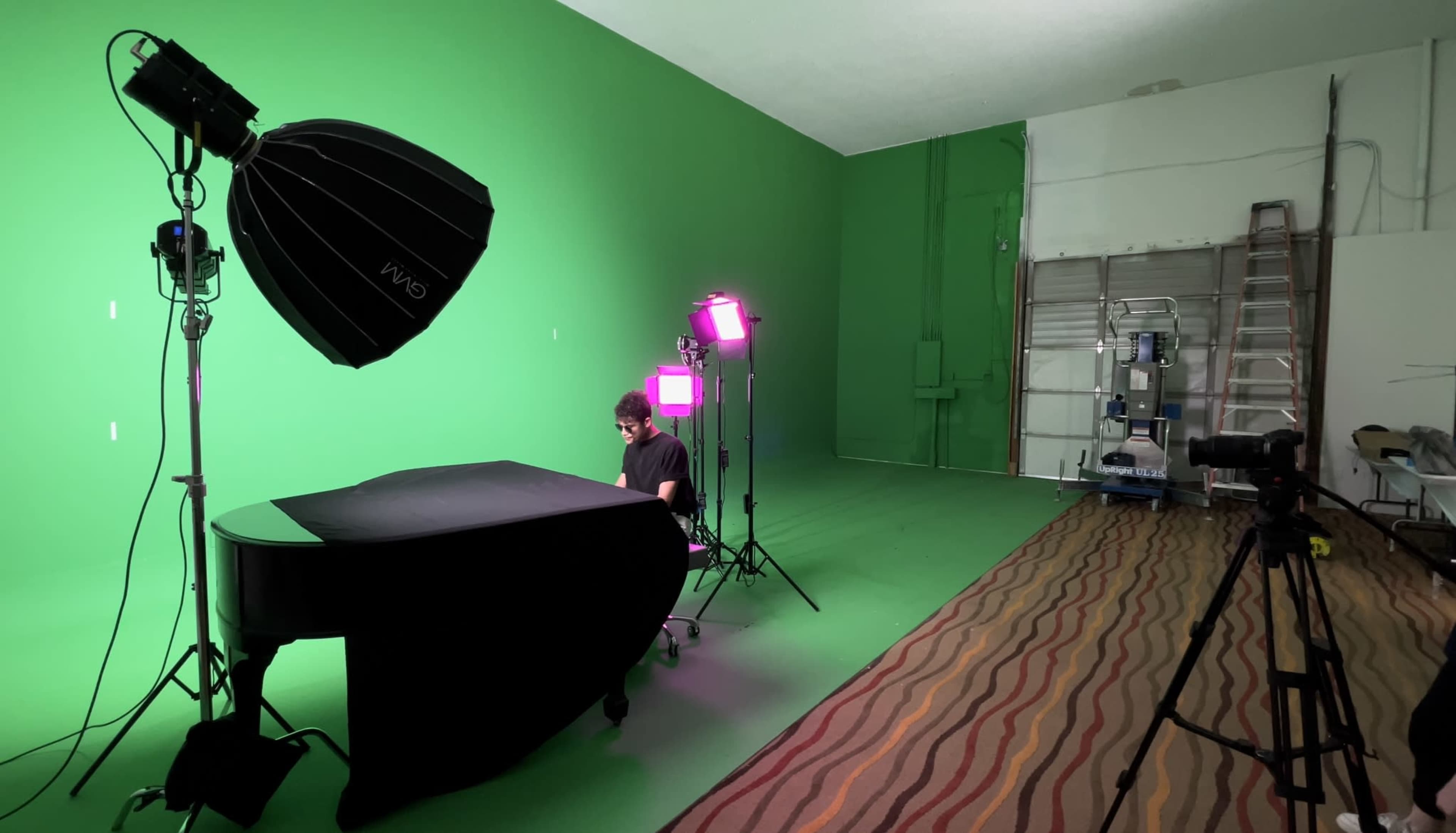 A person sits at a grand piano in a studio set up with green screens, softbox lighting, and additional colored lights.