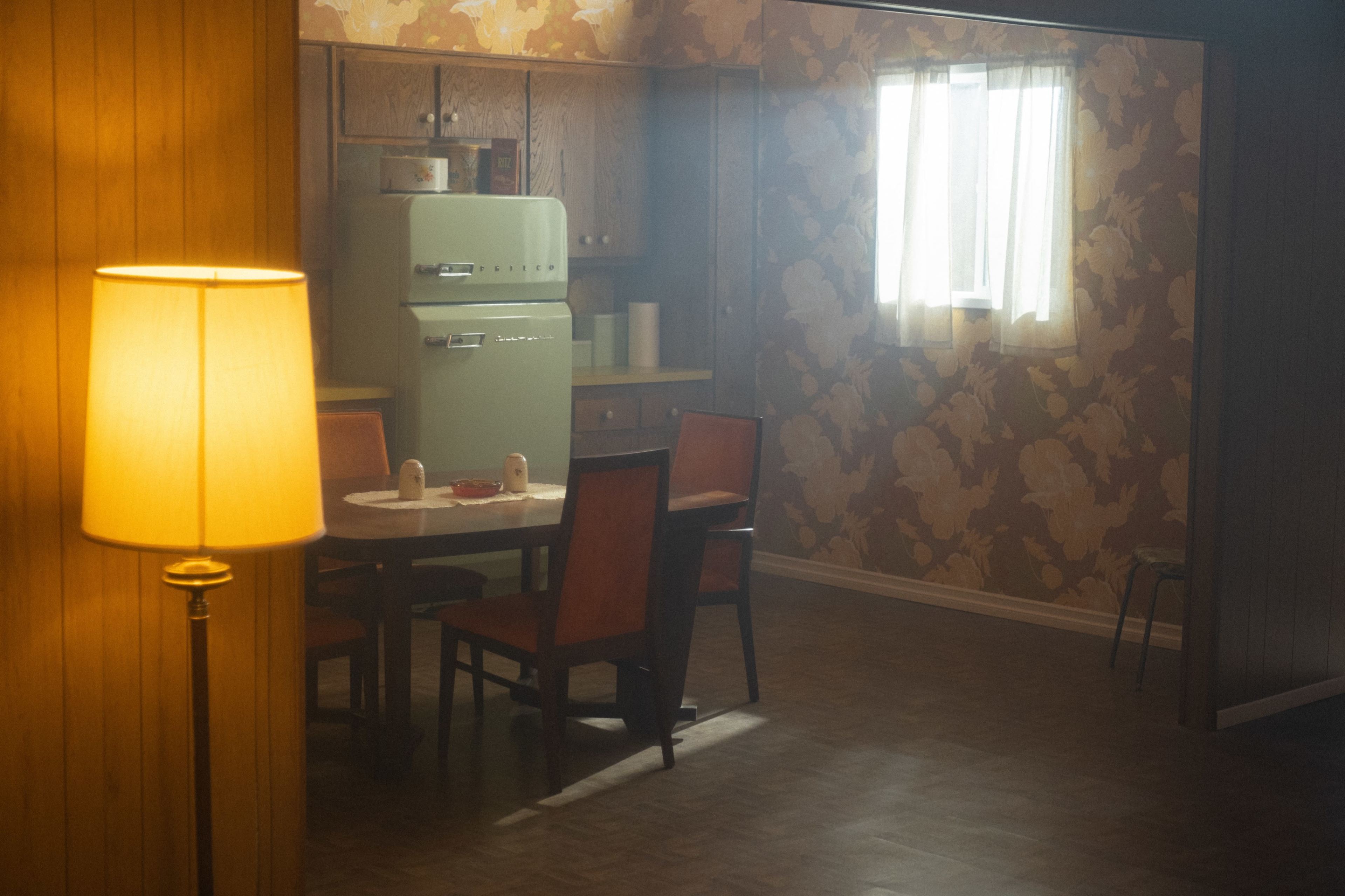 A retro kitchen scene features a table with four orange chairs, a vintage green refrigerator, and floral wallpaper.