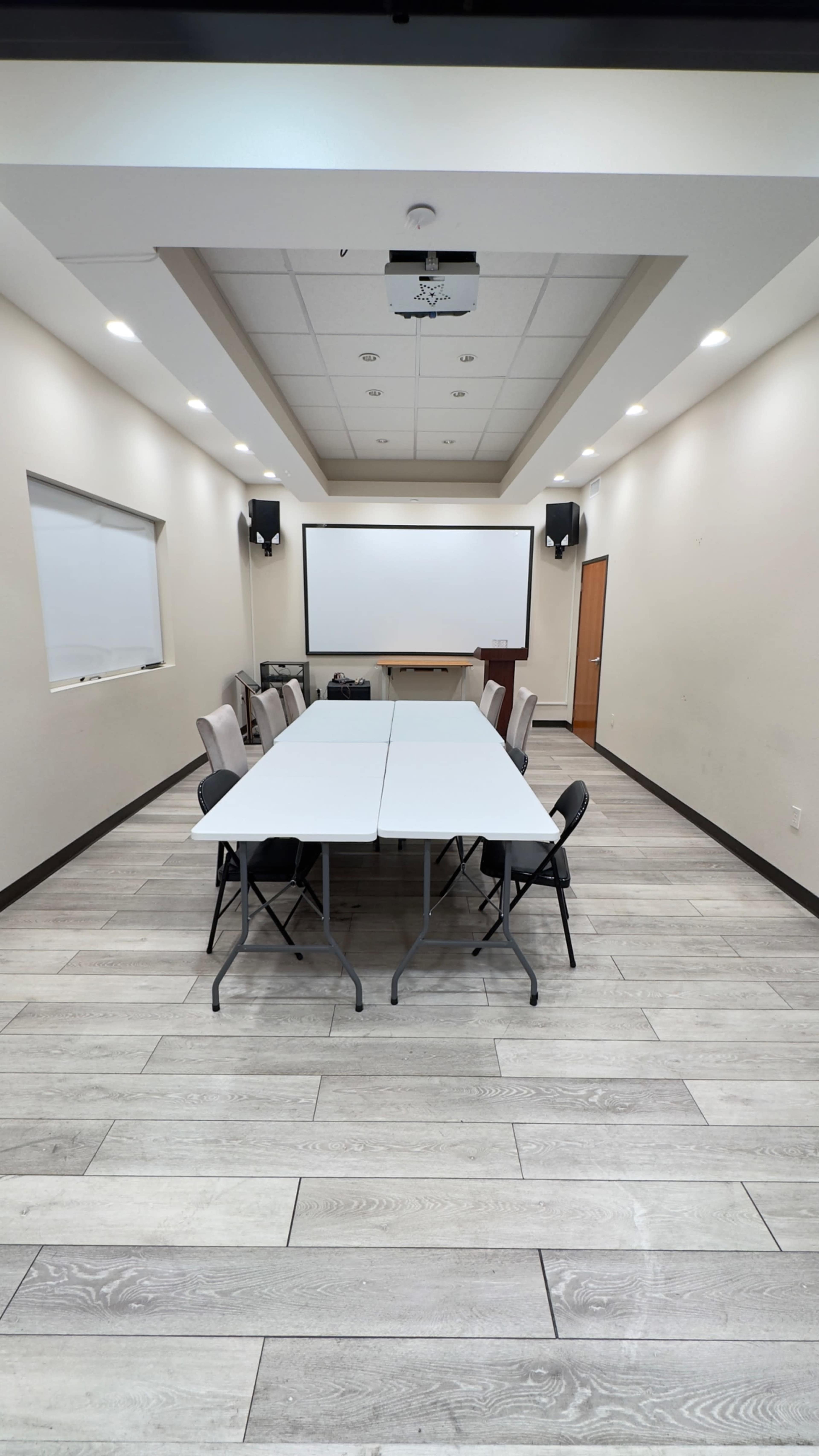 A long table with folding chairs is set up in a conference room featuring a projector screen at one end and windows on the side walls.