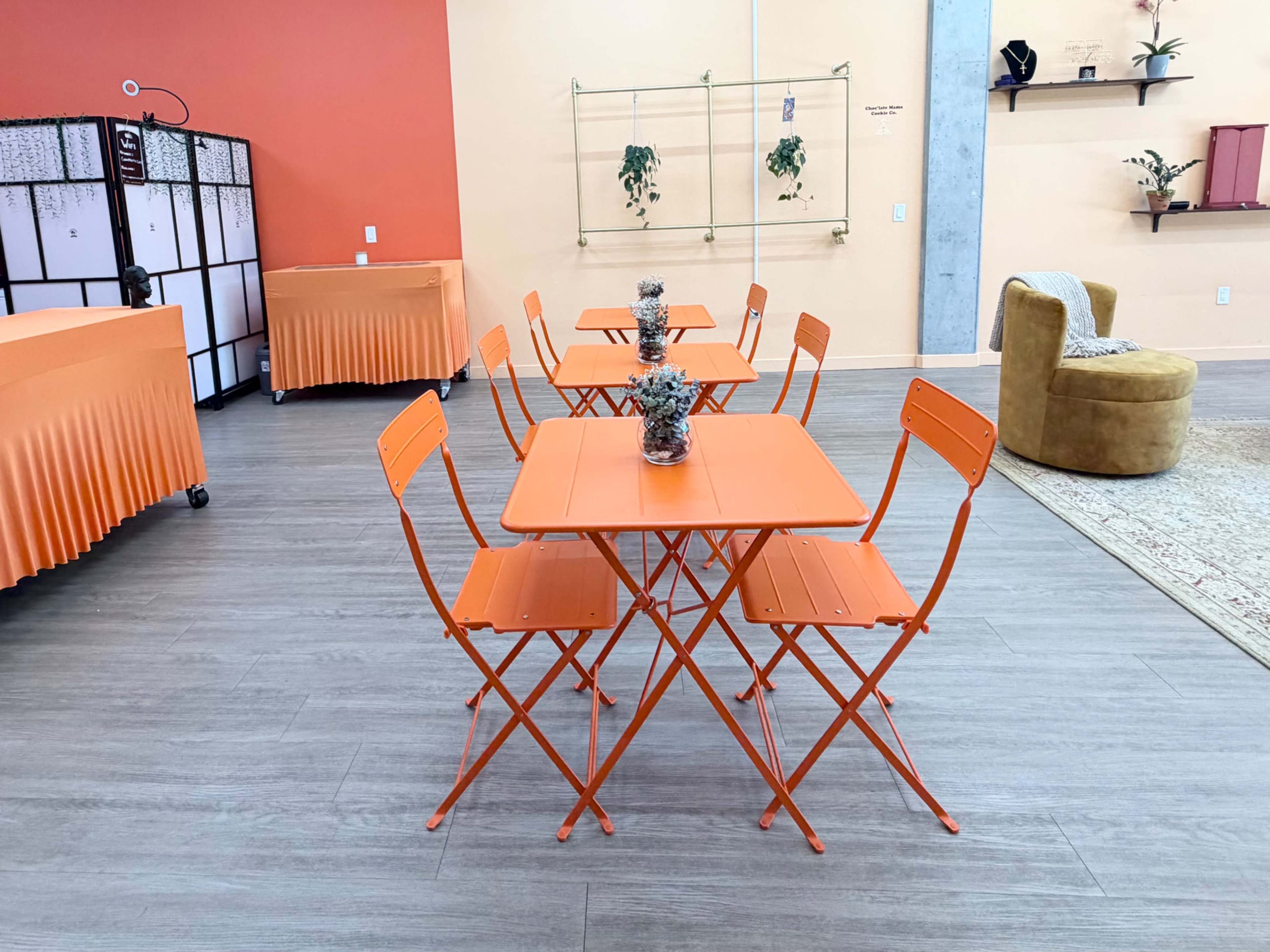 The image shows a bright orange-themed seating area featuring two tables with matching chairs, surrounded by decorative plants and a modern decor style.