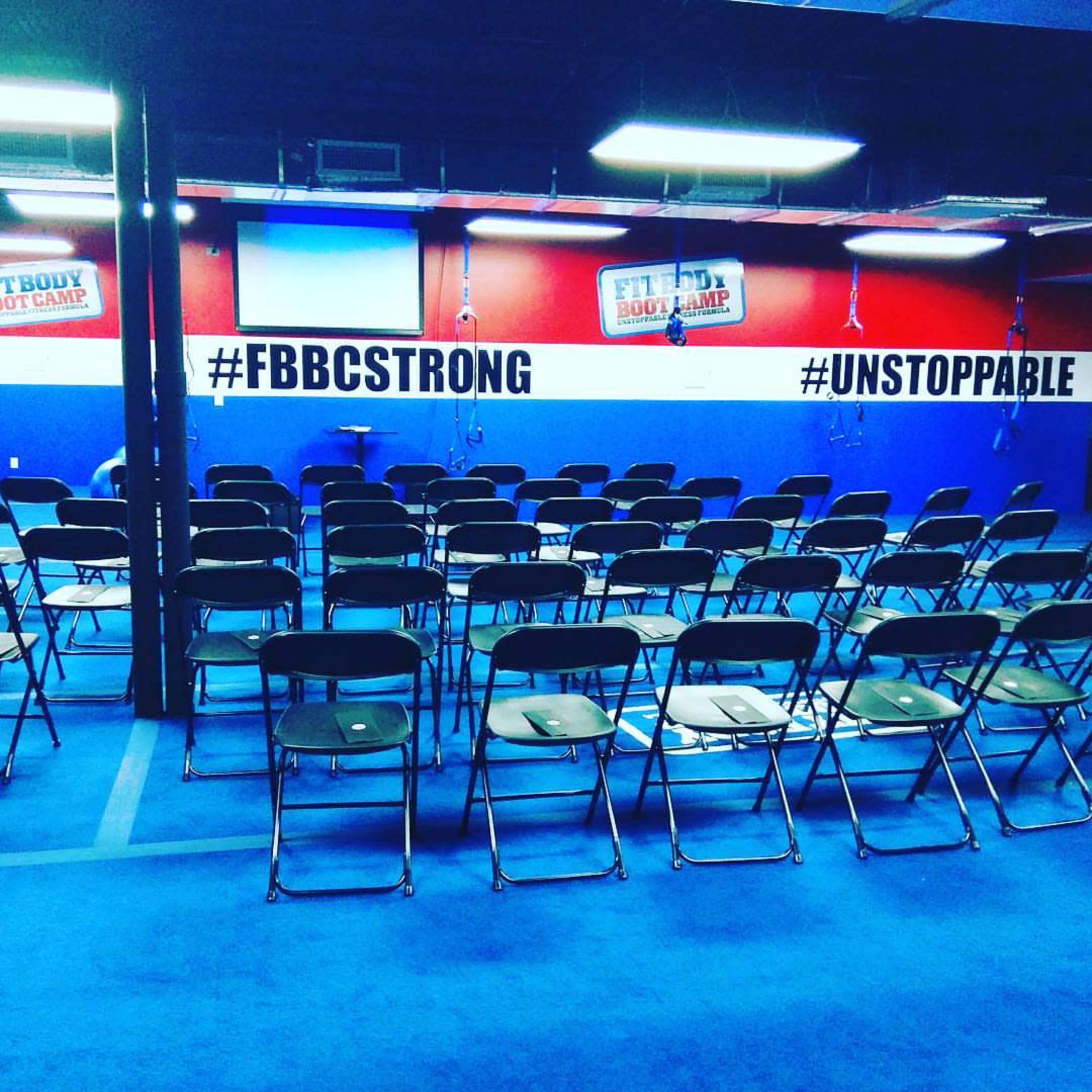 The image shows a fitness studio with rows of empty folding chairs arranged neatly on a blue floor, and motivational hashtags displayed on the walls.