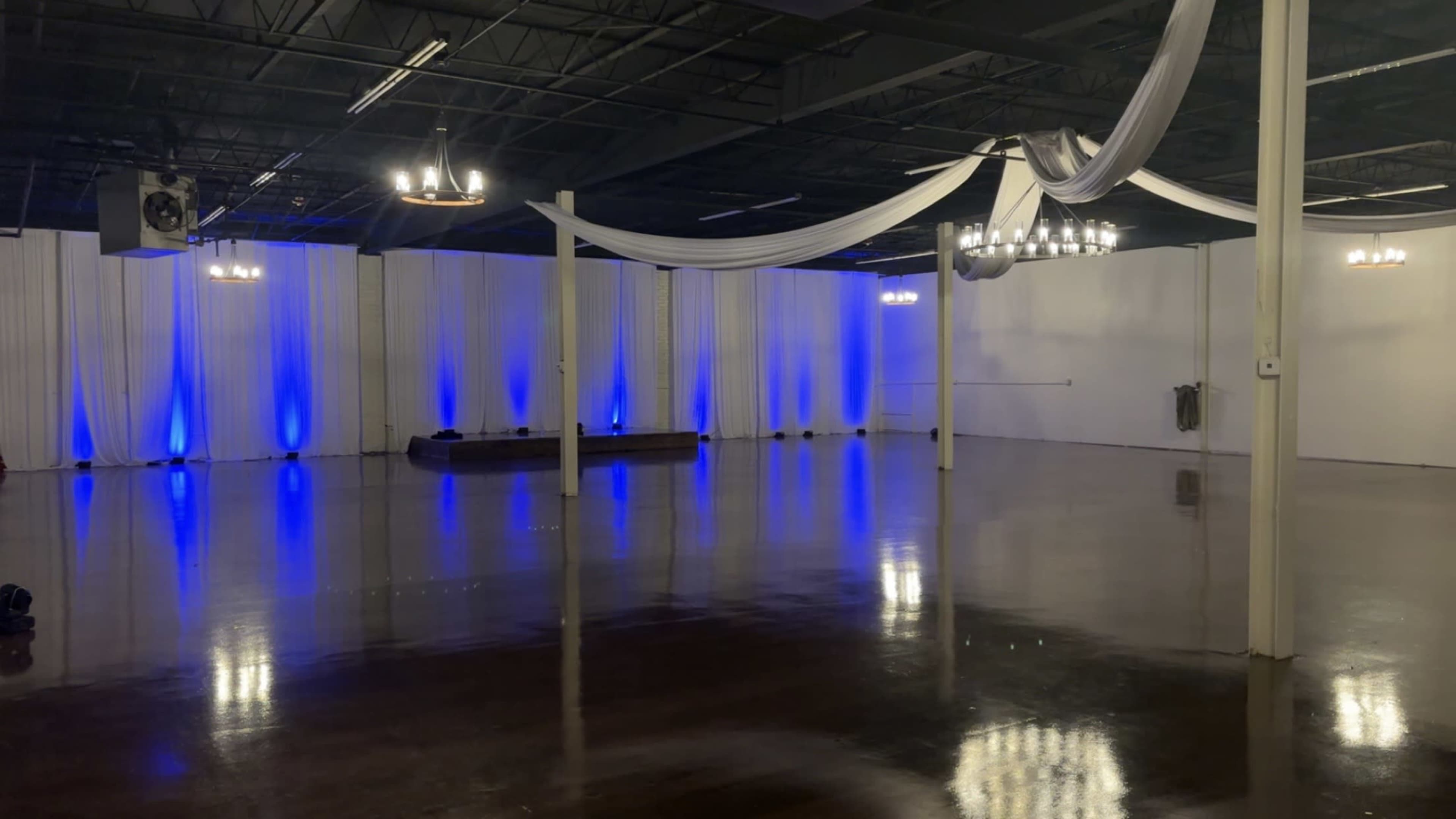 The image shows a spacious event venue with polished floors, draped white fabric, and blue lighting along the walls.