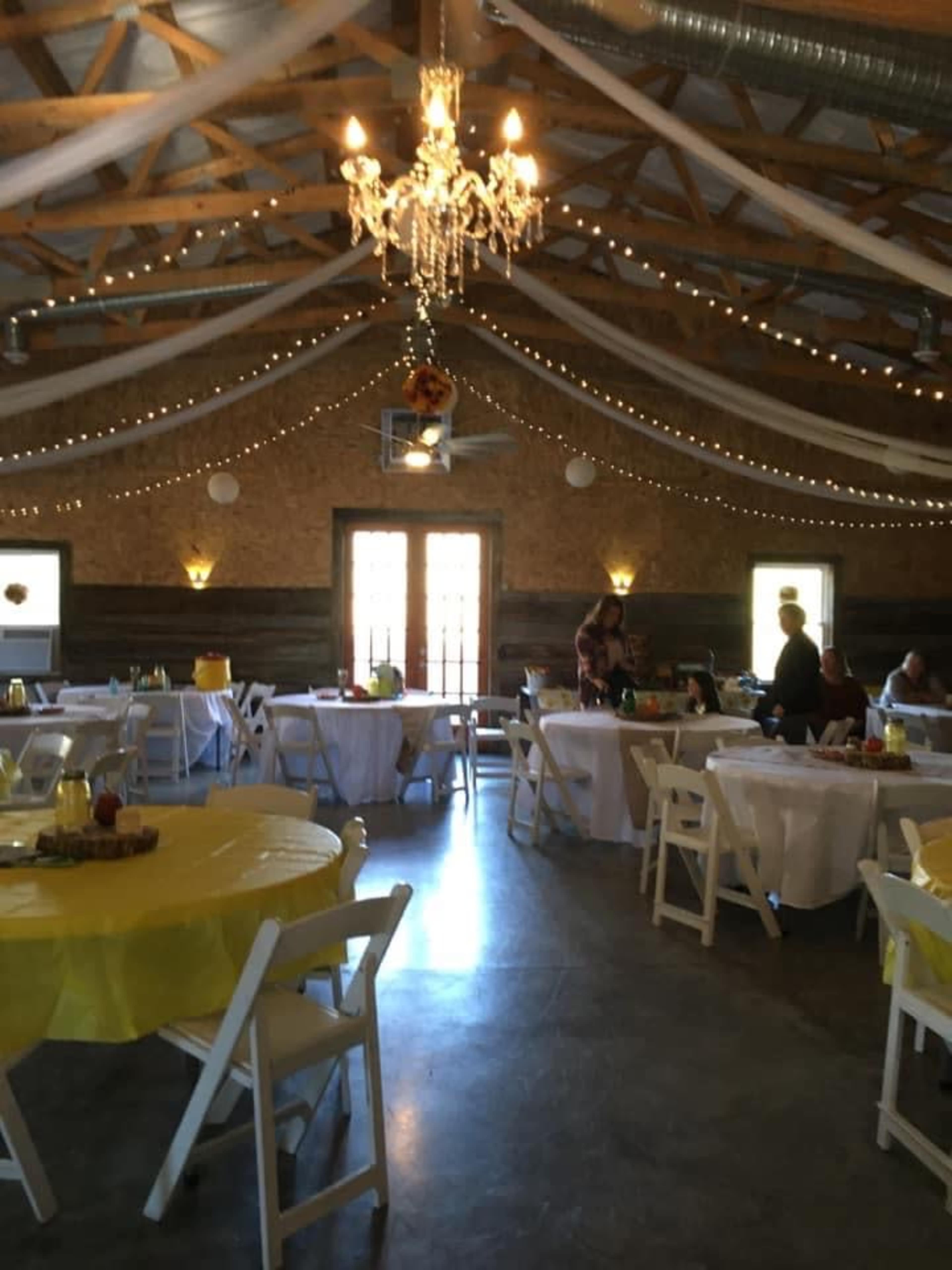 The interior of a banquet hall features round tables covered with white tablecloths, yellow accents, and a chandelier hanging from the ceiling, while guests mingle under string lights and draped fabric.