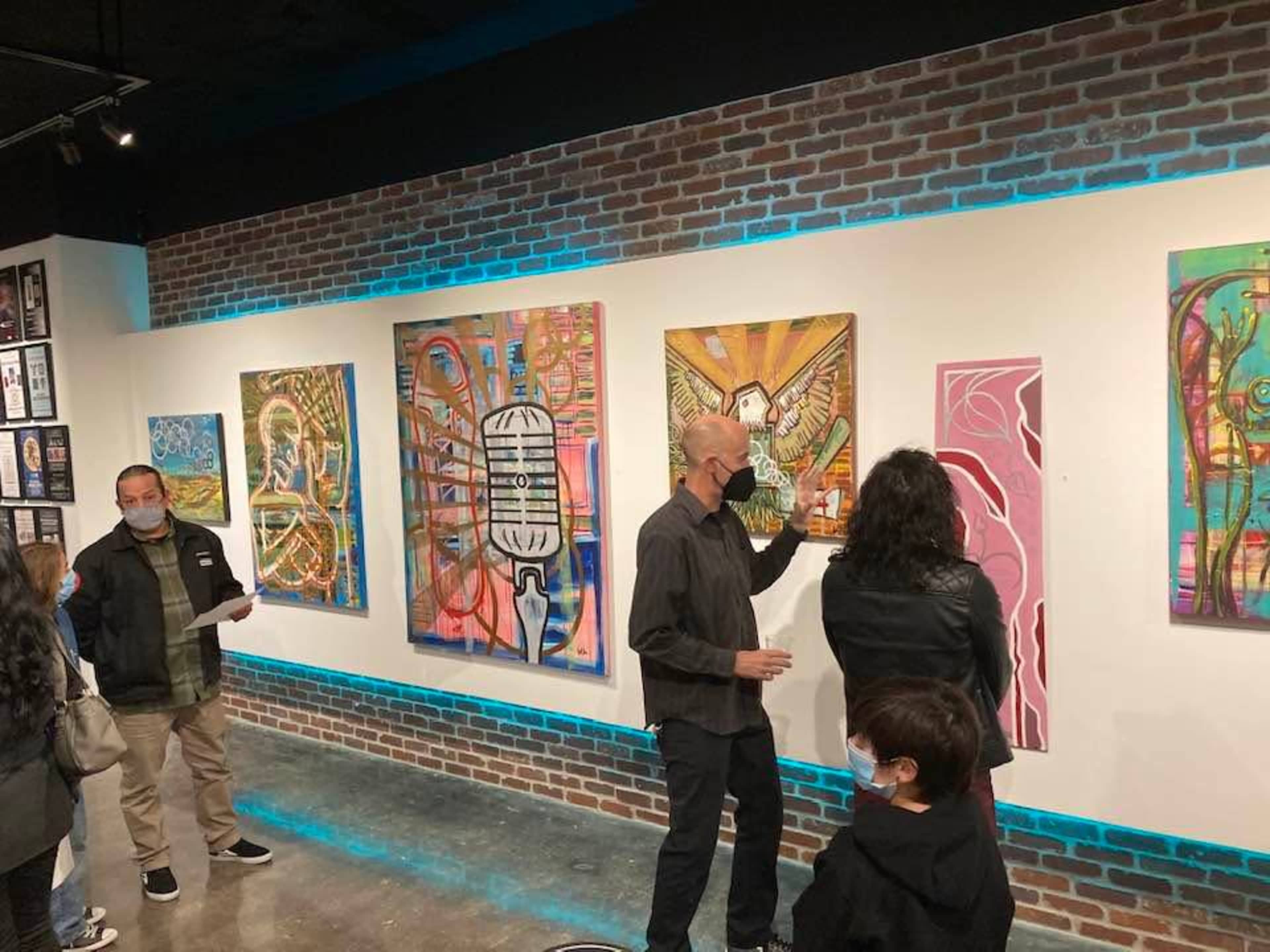 A small group of people examines colorful artwork displayed on a brick wall in an indoor gallery.