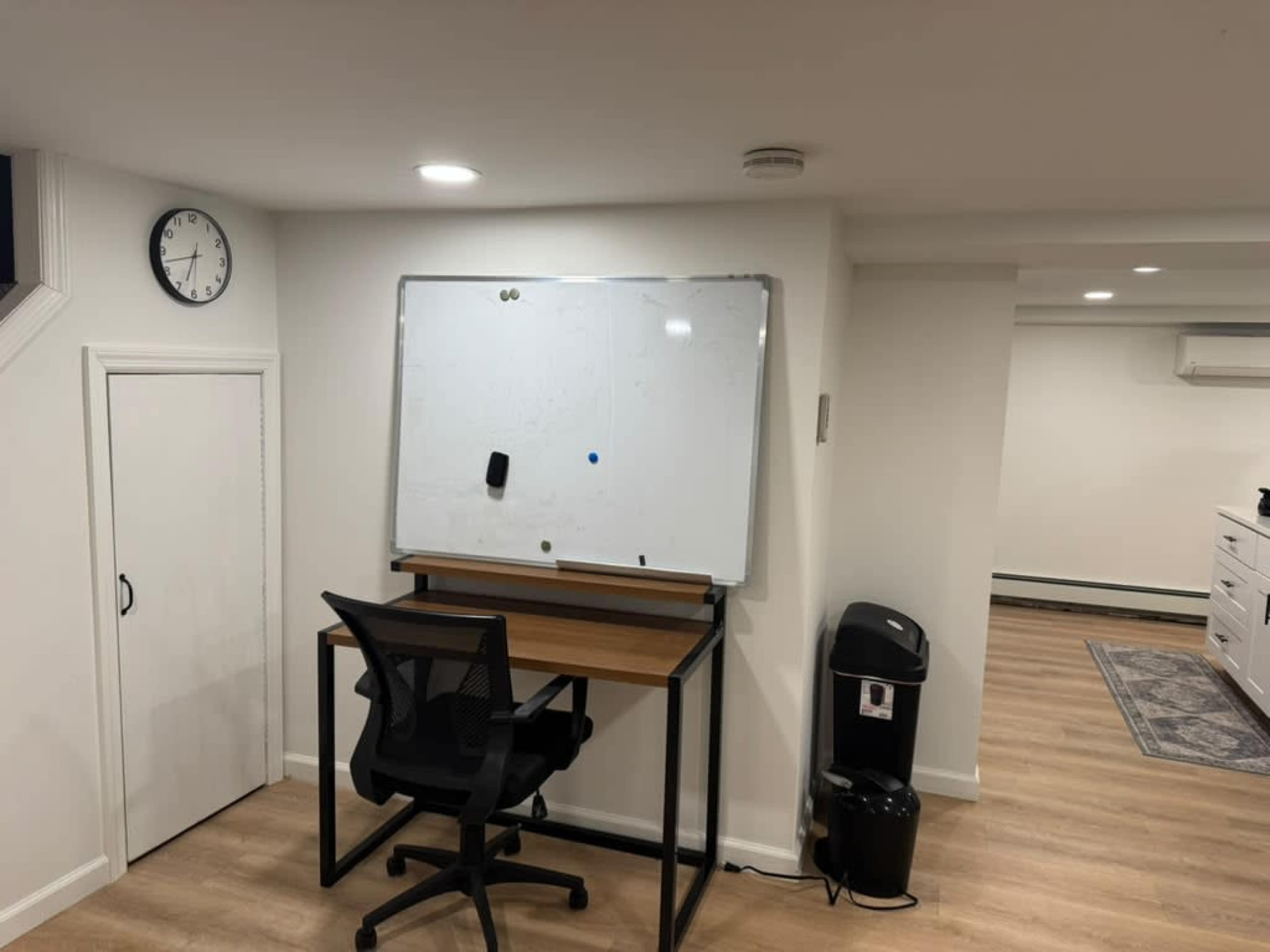 The image shows a minimalistic room with a whiteboard mounted on the wall, a desk with a chair, a clock, and a trash bin, all situated in a space with light wood flooring.