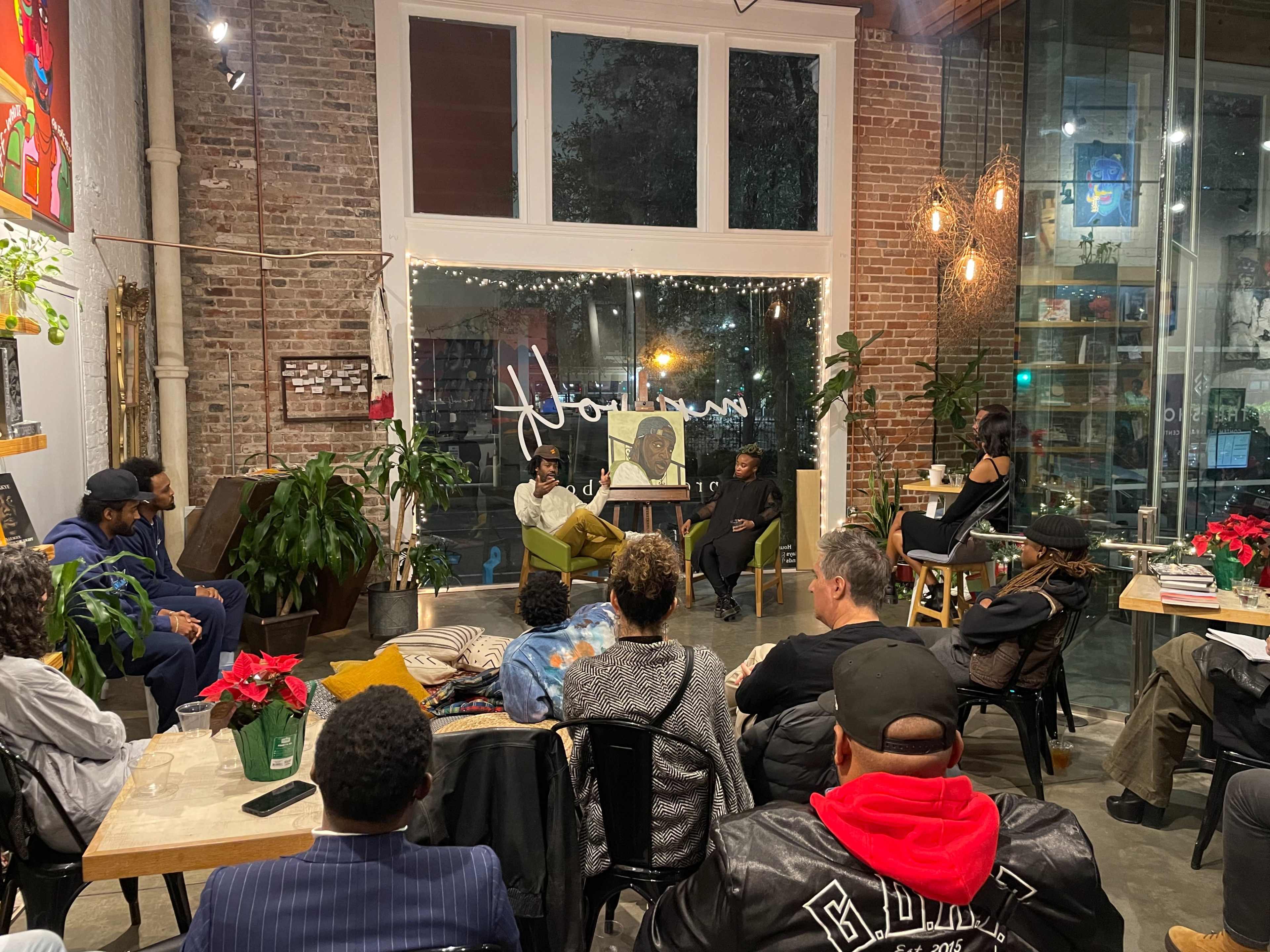 A group of people listen to a speaker in a cozy, brightly lit café decorated with plants and holiday flowers.