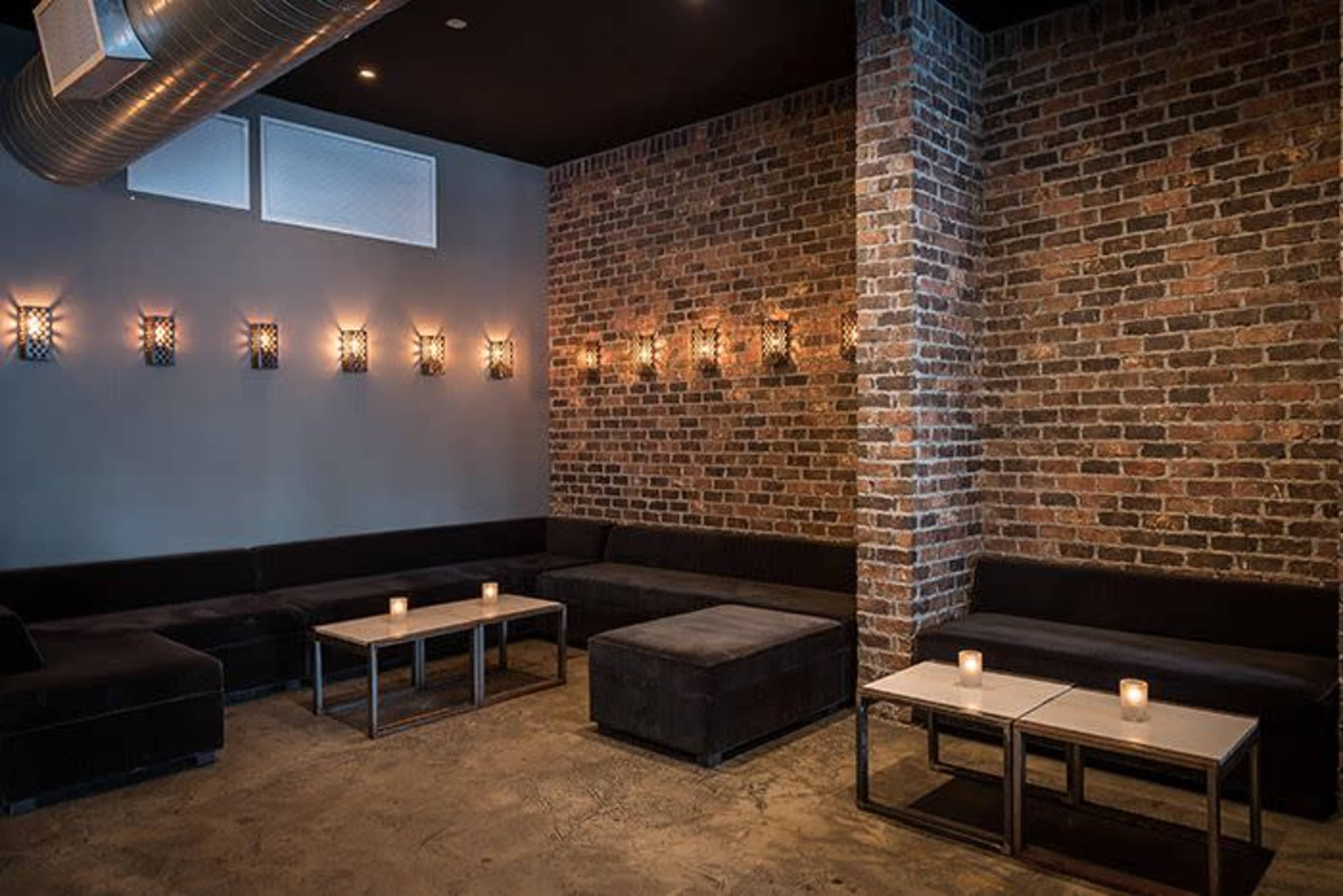 The scene shows a dimly lit lounge area with exposed brick walls, low black seating, and metal tables topped with lit candles.