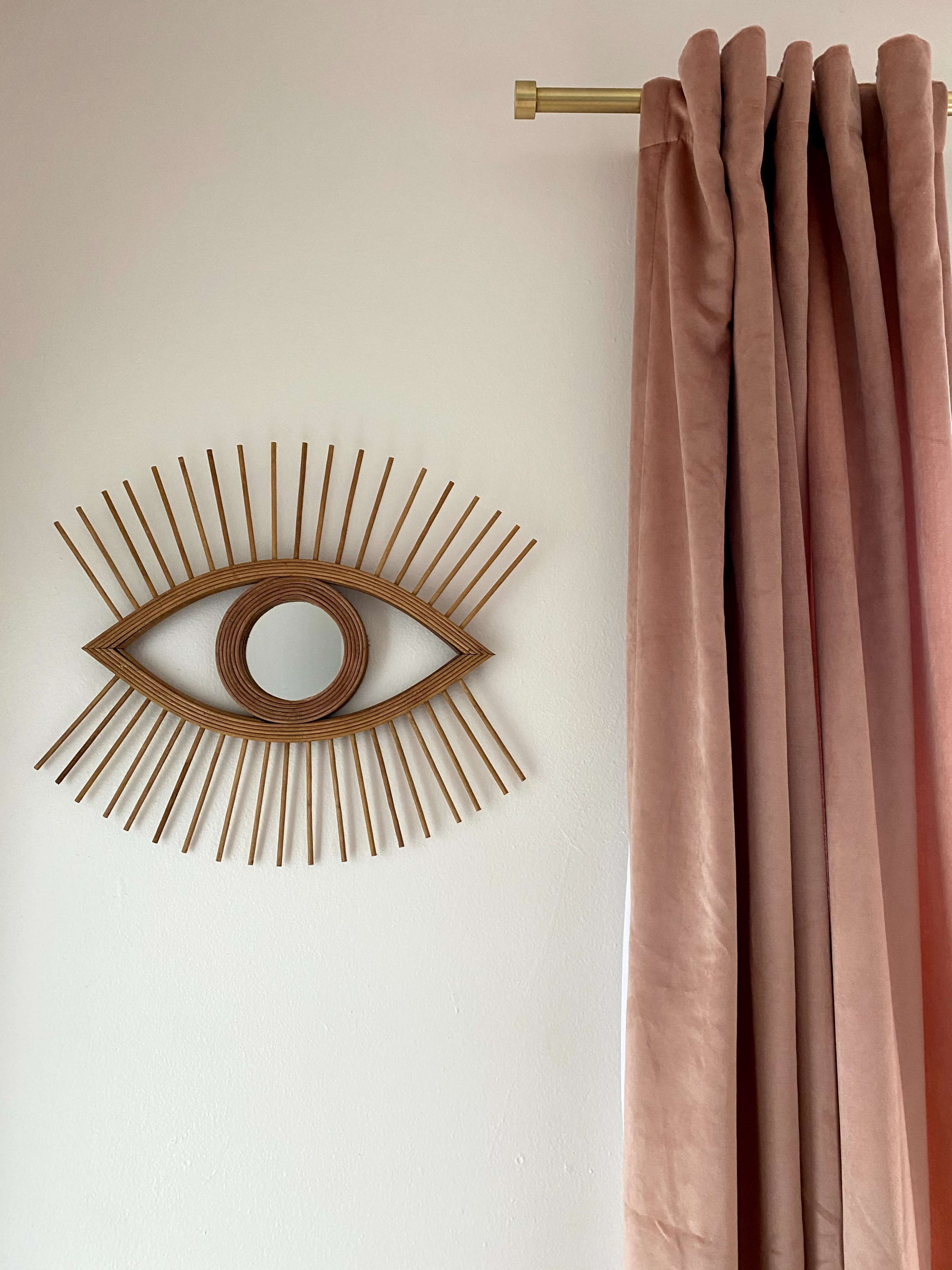 A decorative wooden eye-shaped wall art beside a pink velvet curtain hanging from a brass rod.