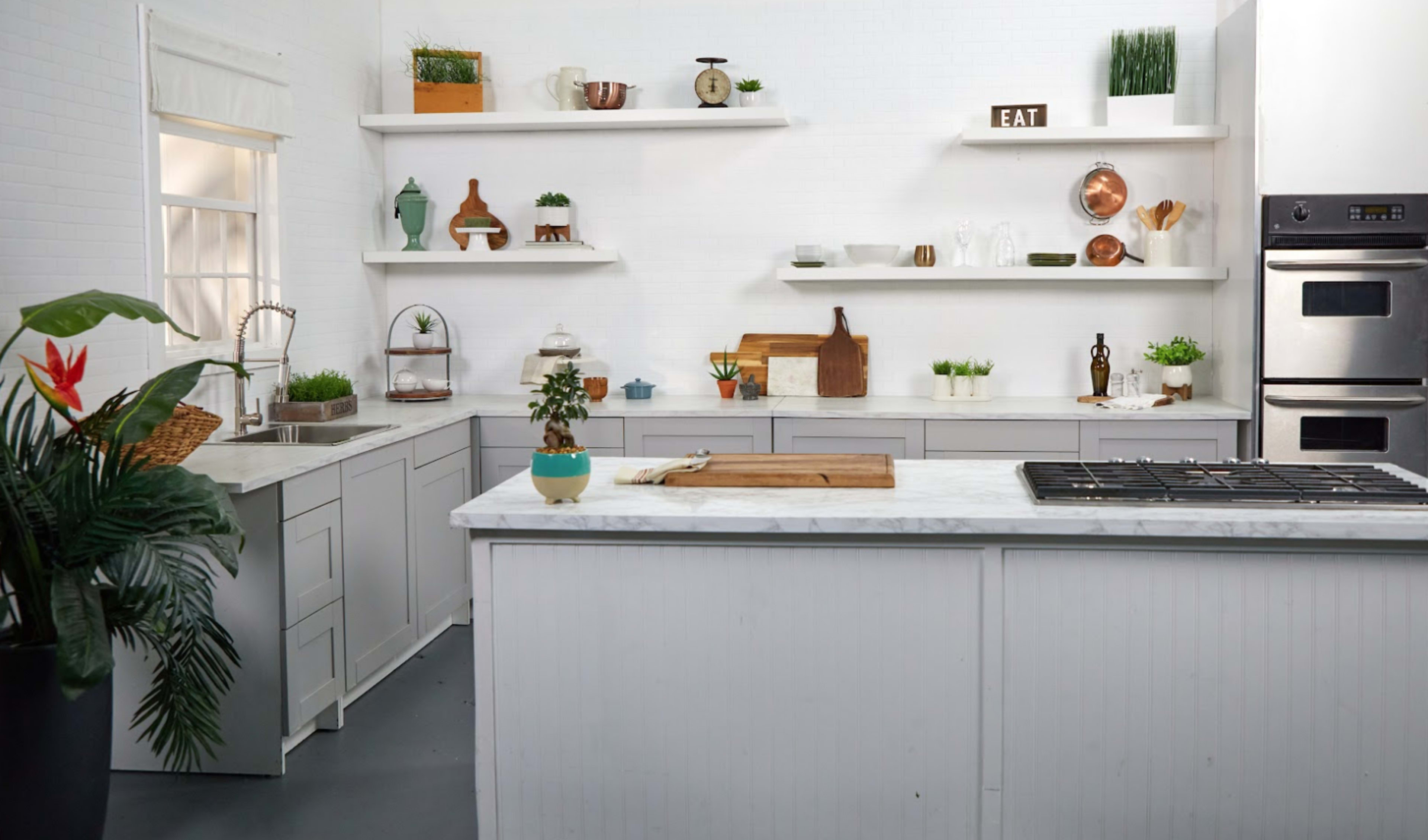 A modern kitchen features gray cabinetry, a marble countertop island, and open shelves with decorative items and plants.