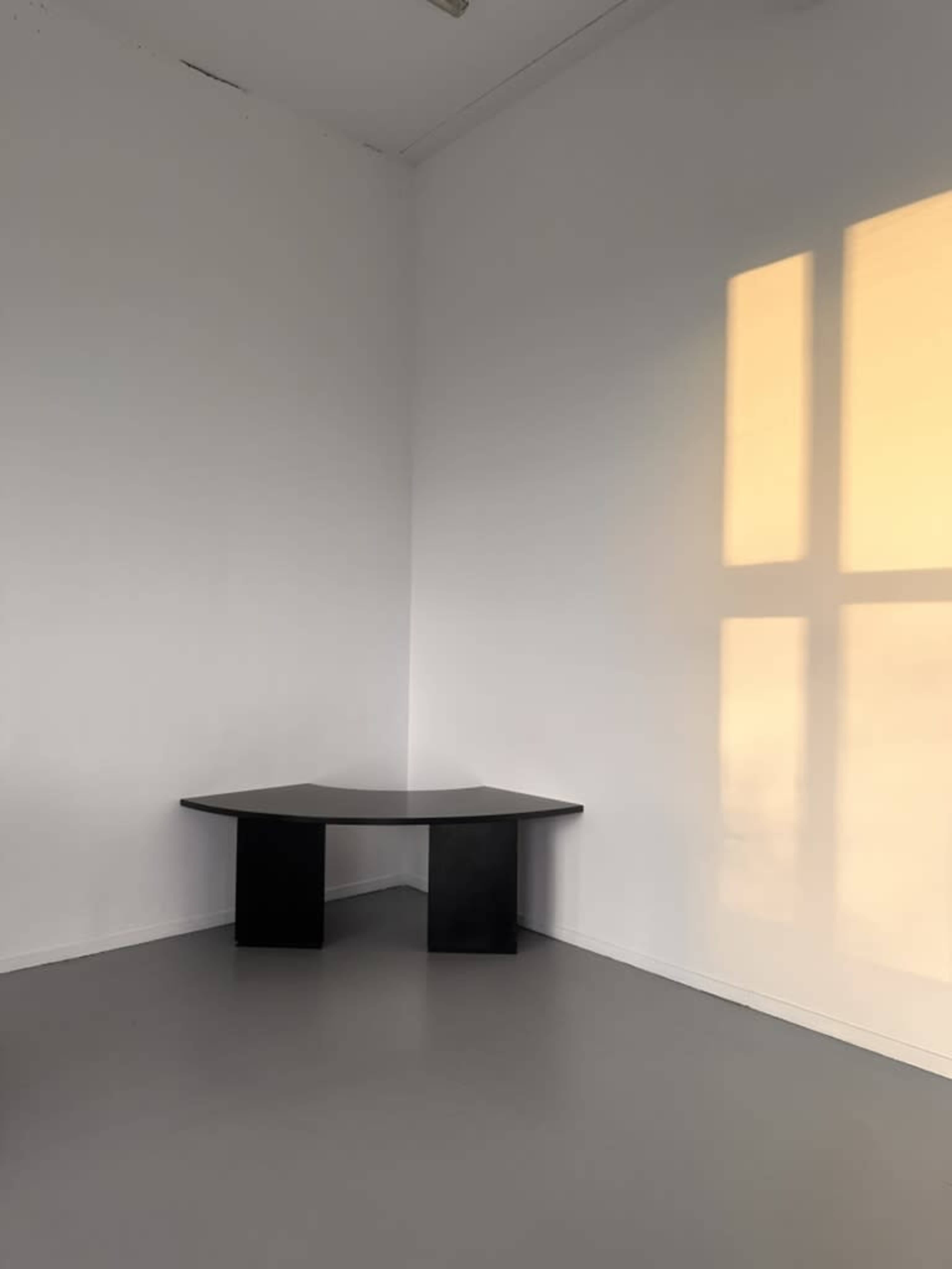 A black, corner table is positioned in an empty room with white walls and gray flooring, illuminated by soft light casting a shadow on the wall.