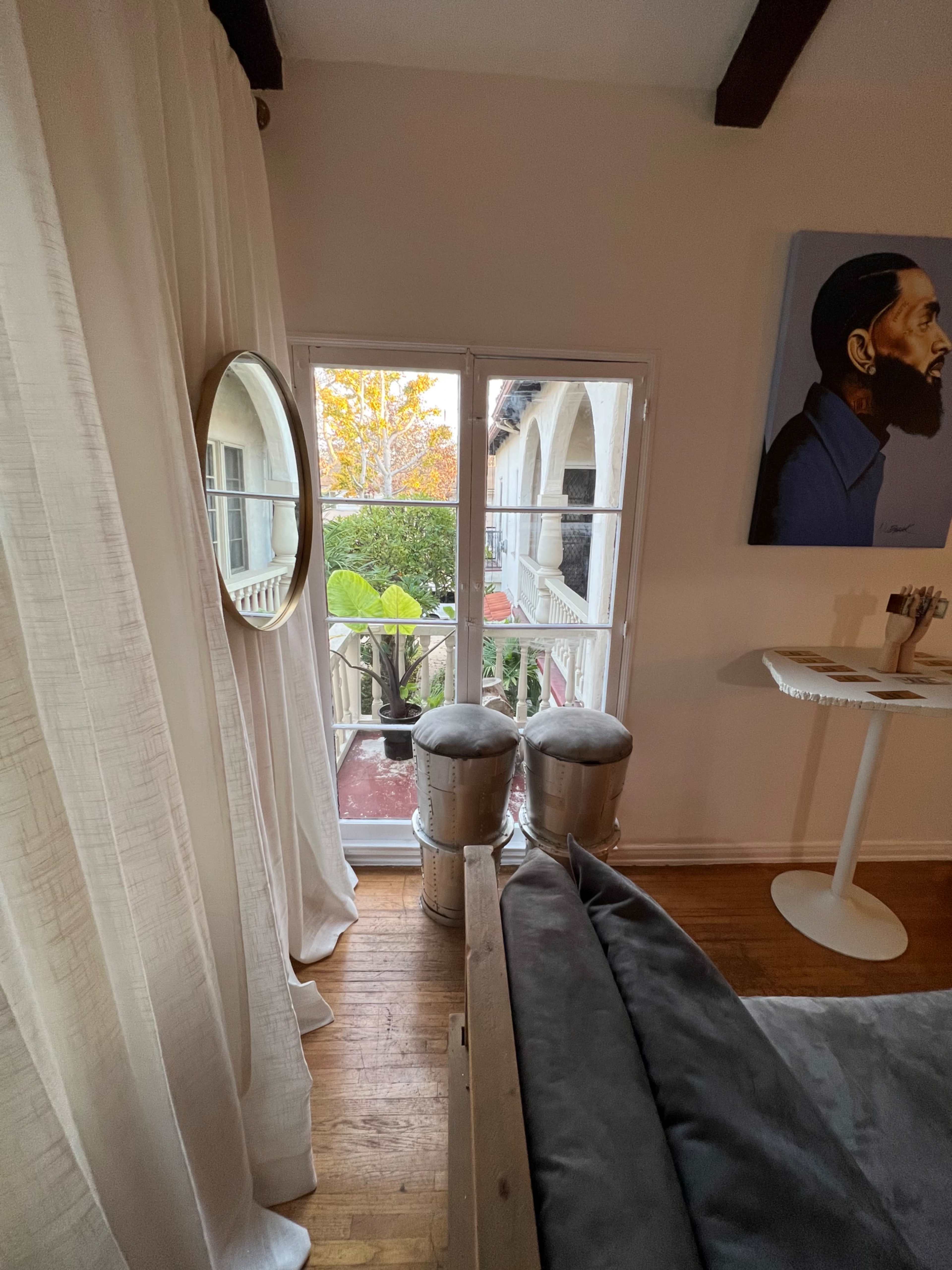 A bedroom features a large mirror and two stools near a window that opens to an outdoor terrace with visible greenery.