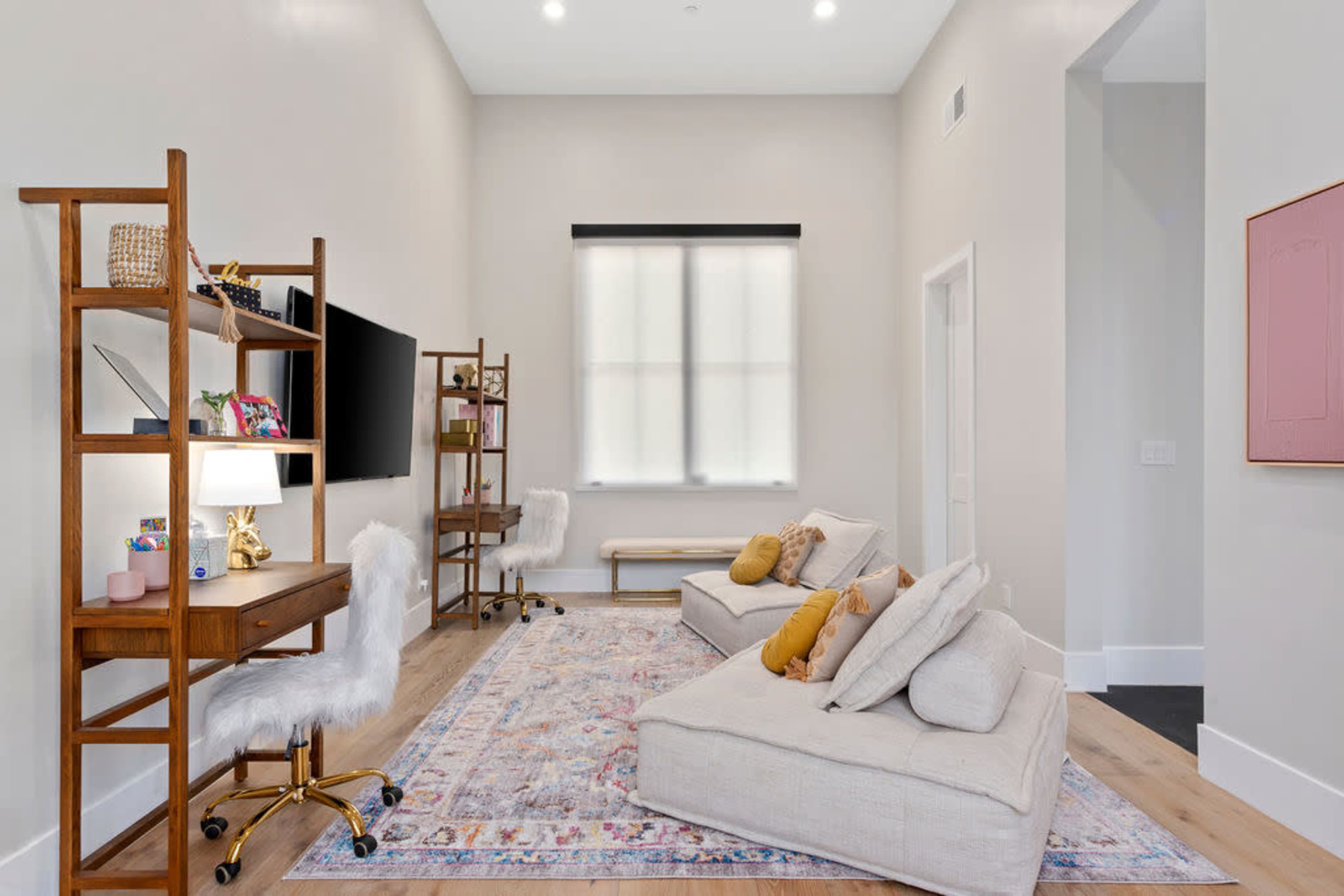 A well-lit modern room features a low seating area with a large rug, a desk with a lamp, and a wall-mounted television, complemented by a bench and decorative shelving.