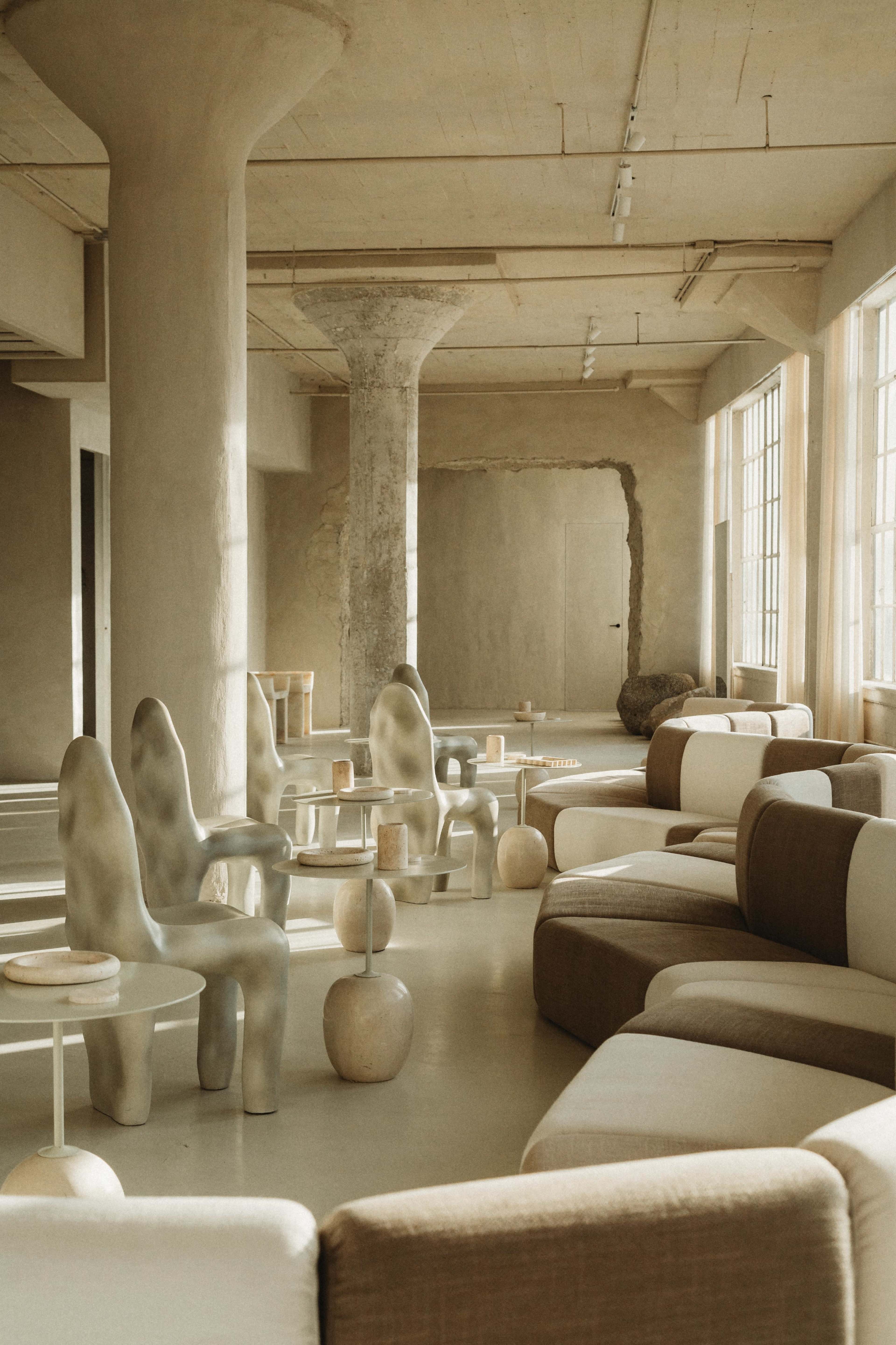 A spacious interior features sculptural chairs and minimalist furniture arranged around coffee tables, with large windows allowing natural light to fill the room.