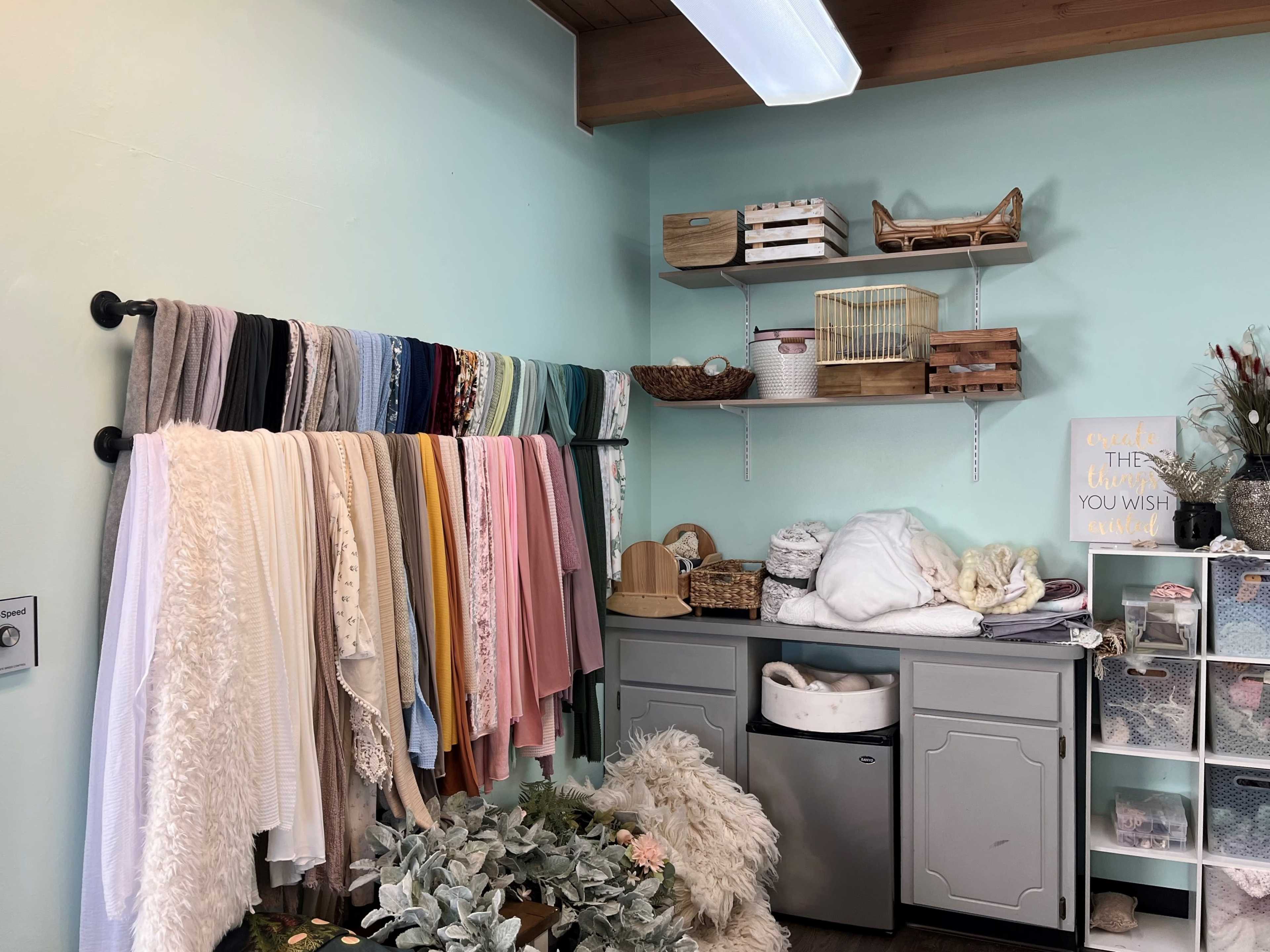 The image shows a neatly organized room with colorful fabric rolls hanging on a wall, shelves filled with decorative boxes, and a cabinet containing various textiles and a plant arrangement.