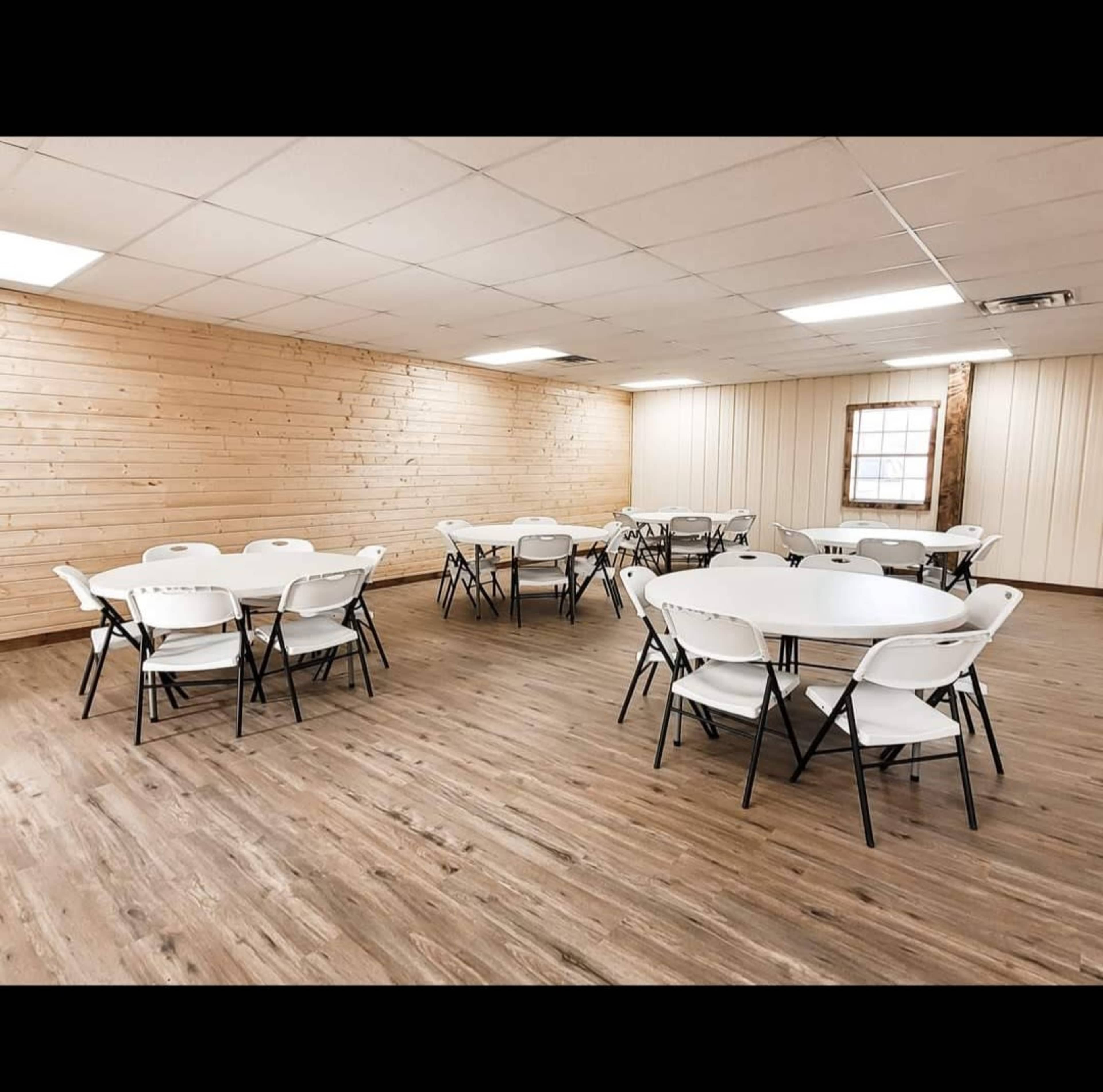 A spacious room features several round tables surrounded by folding chairs, with a wooden wall and vinyl flooring.