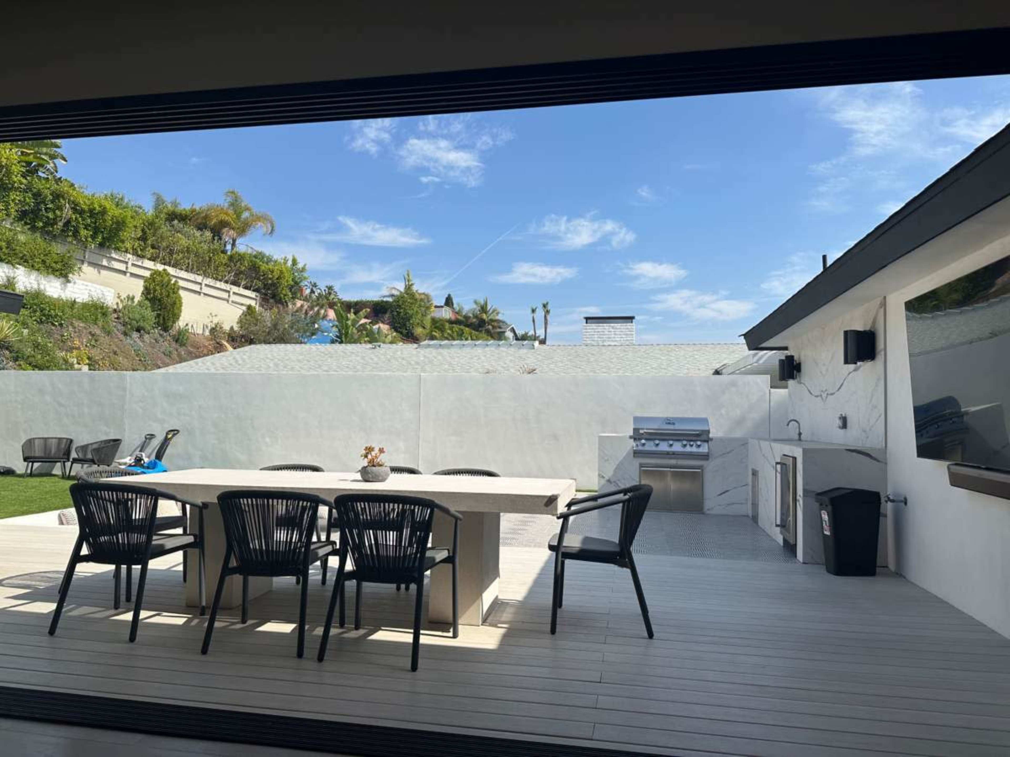 The image shows a spacious outdoor patio with a dining table and chairs, a barbecue grill, and a lawn area under a clear blue sky.