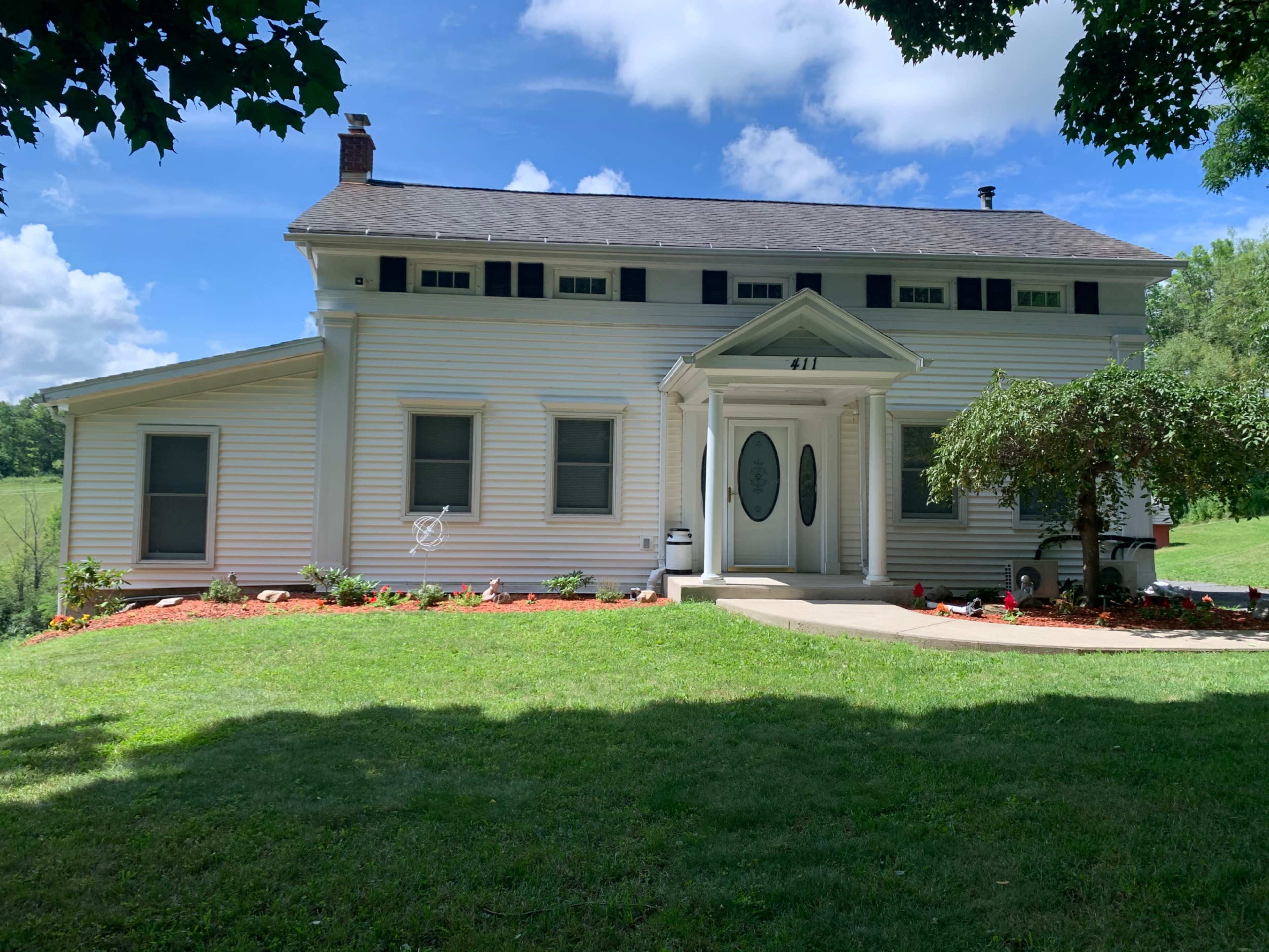 A two-story white house with a front porch, surrounded by a green lawn and flower beds.
