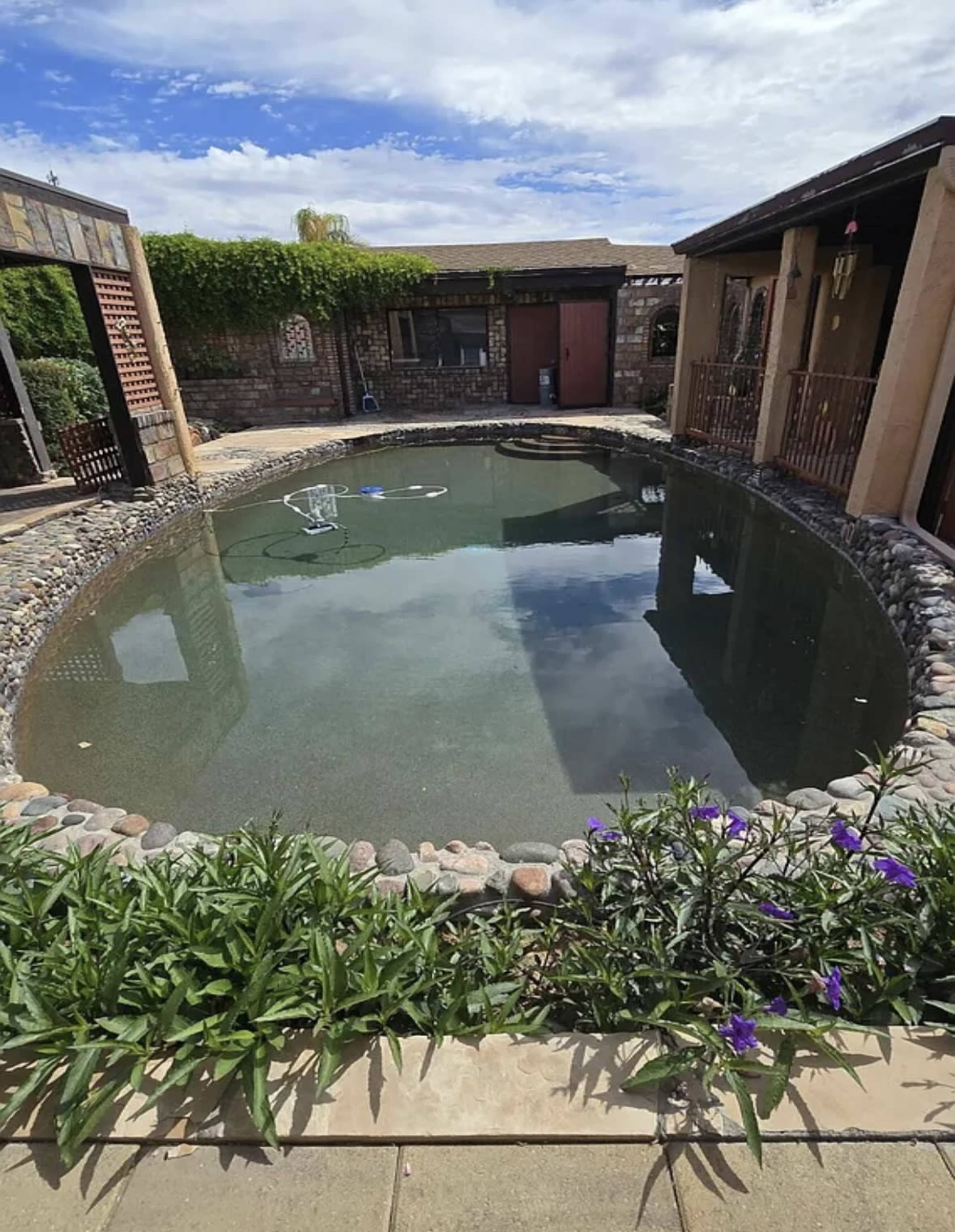 The image shows a circular, outdoor swimming pool surrounded by stone and brick structures, with greenery and purple flowers along the edge.