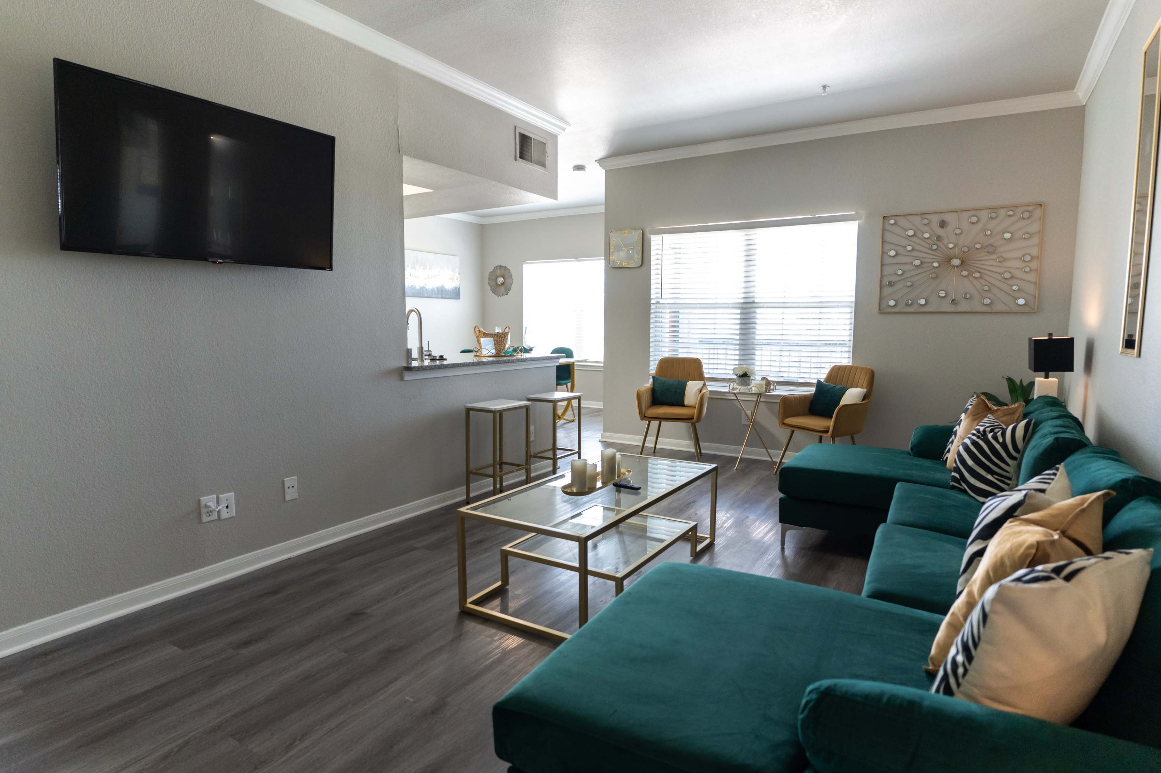 The living room features a green sectional sofa, a glass coffee table, two chairs, and a television mounted on the wall.