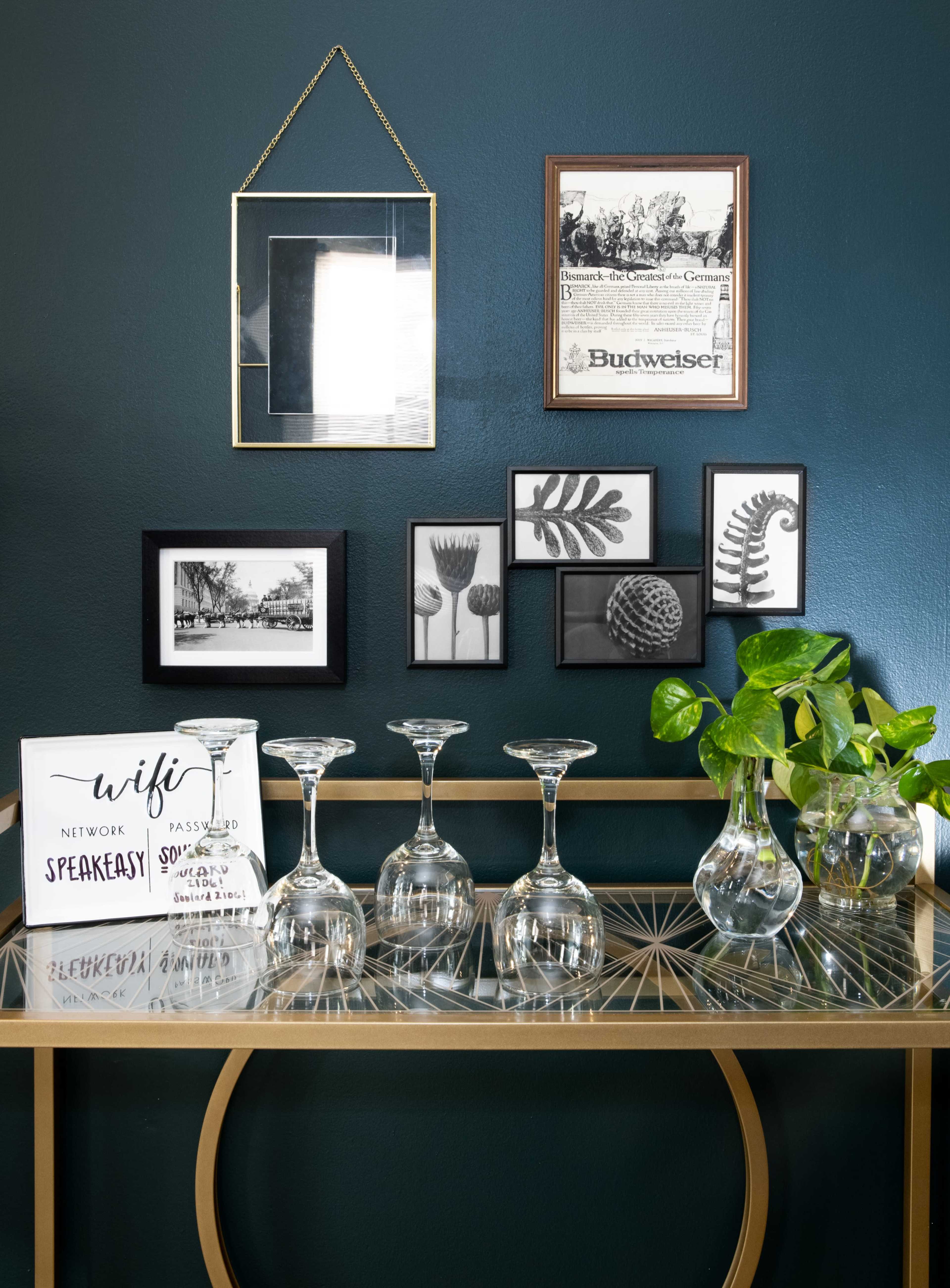 A console table displays several crystal wine glasses, a potted plant, and various framed artworks against a dark green wall.